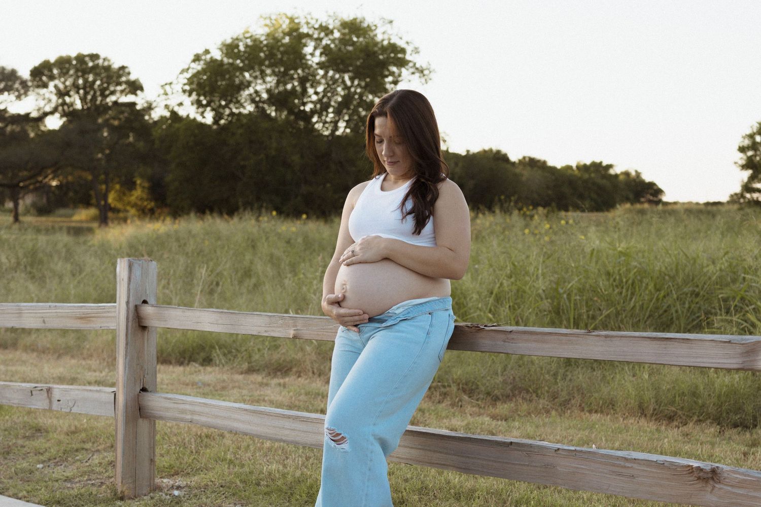 A pregnant mom in light blue pants cradles their pregnant belly while standing by a wooden fence at sunset in a rural setting.