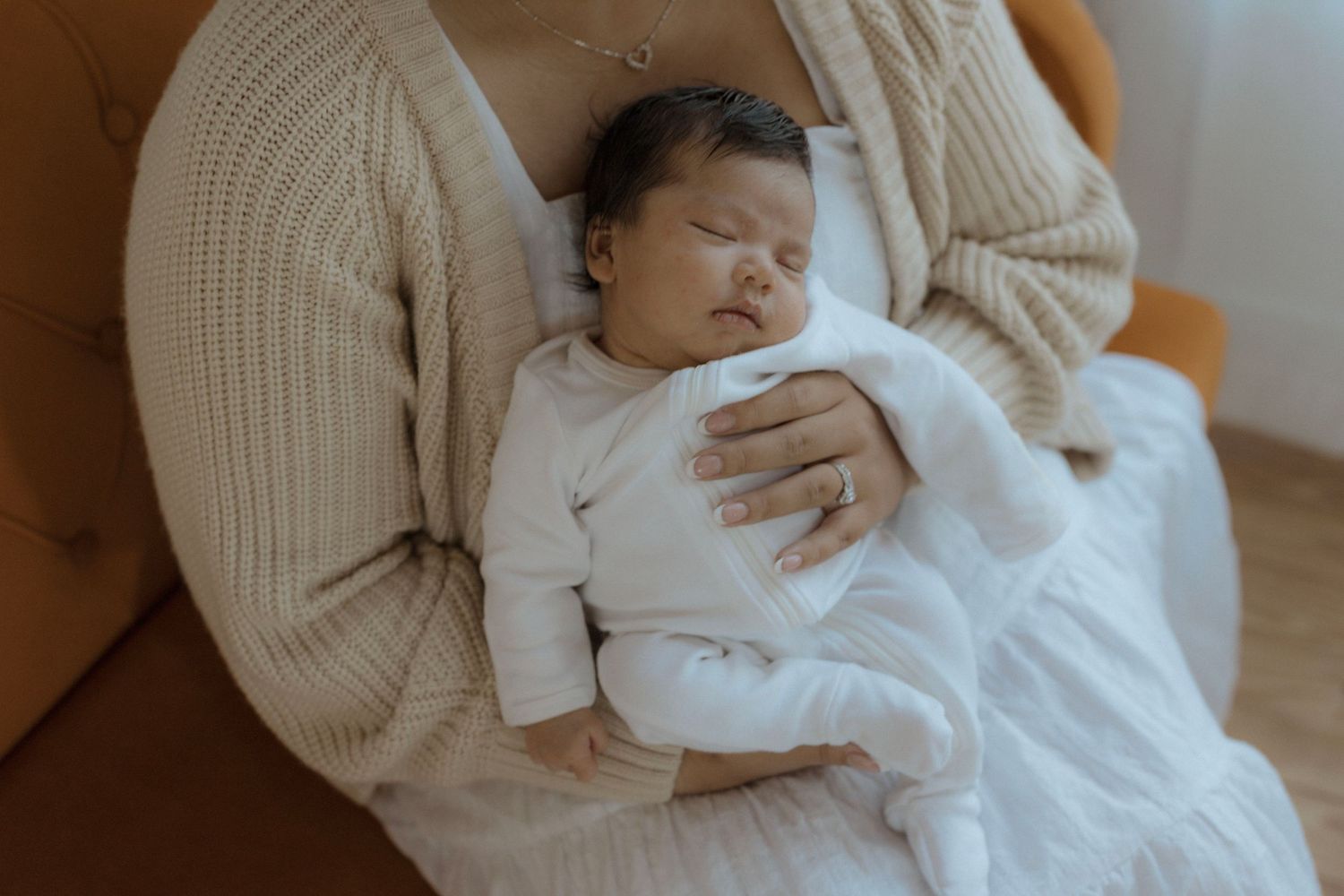 Sleeping newborn baby wearing white onesie rests peacefully in caregiver's lap on beige chair.