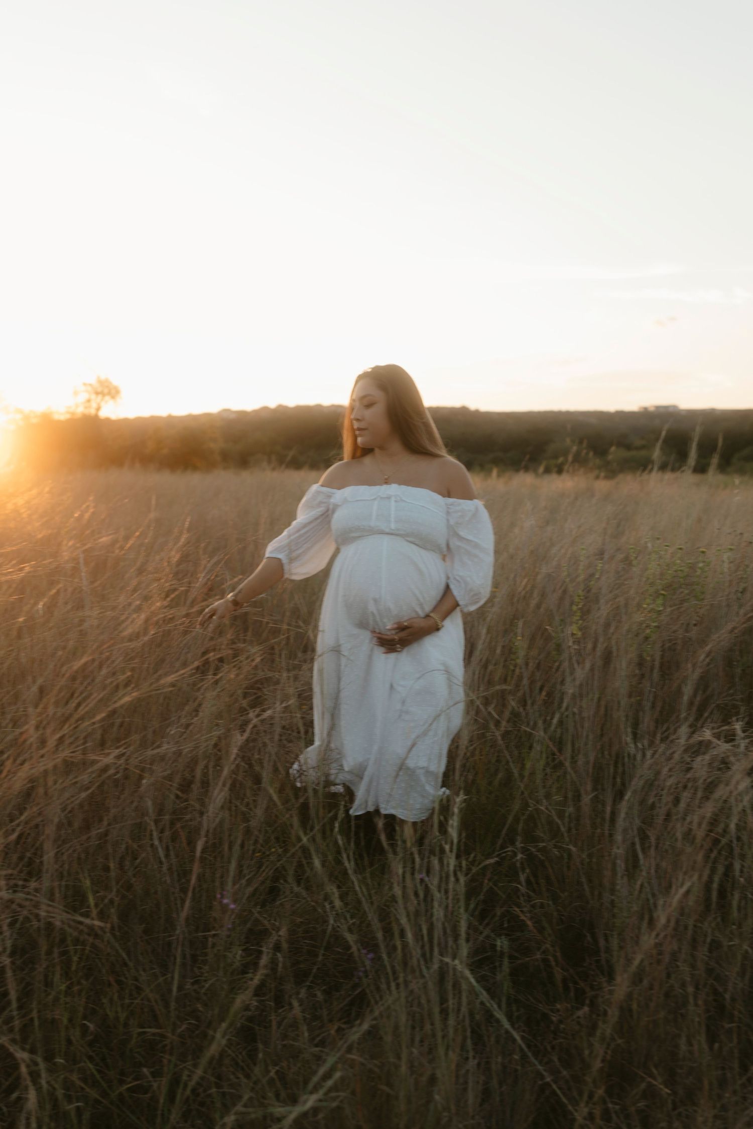 A serene maternity portrait of a woman in white dress touching her belly during sunset in a field.