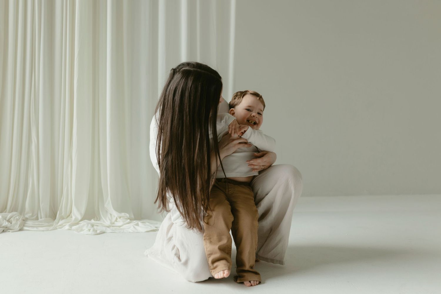 Documentary motherhood photo of a mom tickling her son during a minimalist white studio session.