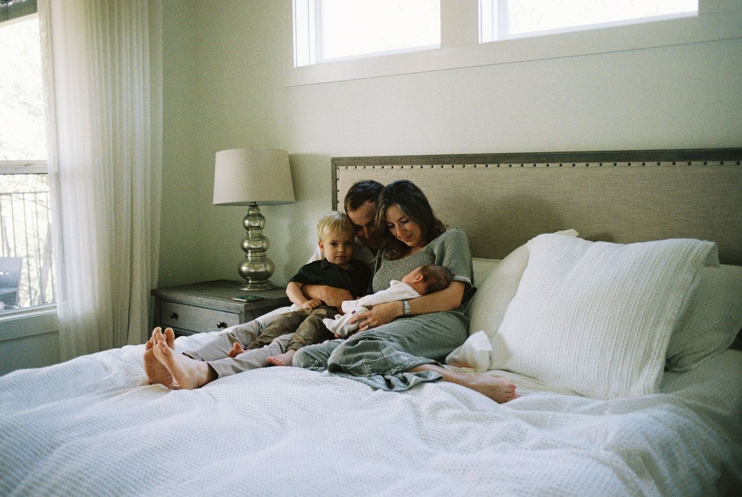 A cozy morning scene with people relaxing together on a white bed while looking at something in a sunlit bedroom.