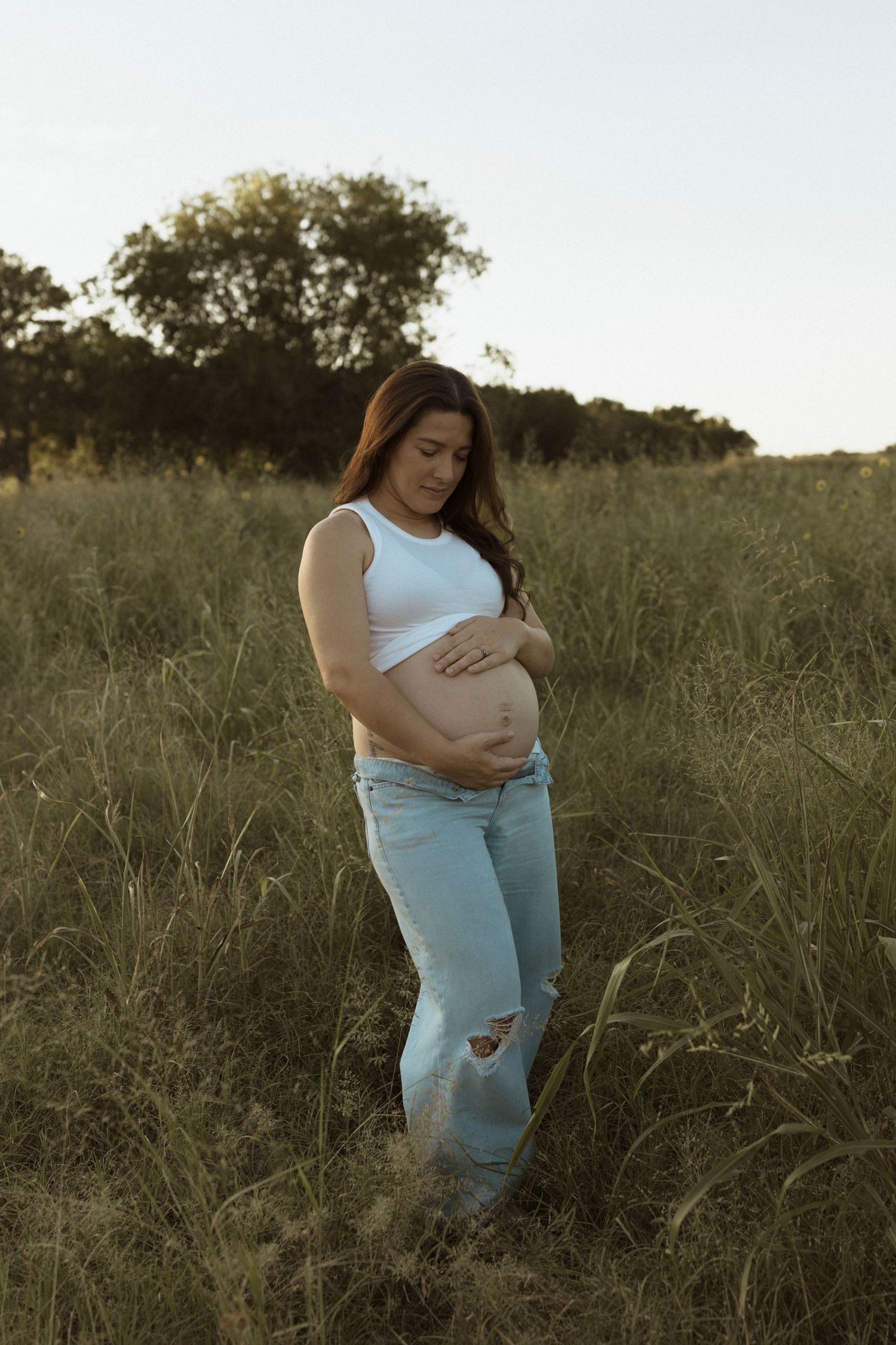 A pregnant mom in jeans and white top stands thoughtfully in a golden grassy field at sunset.