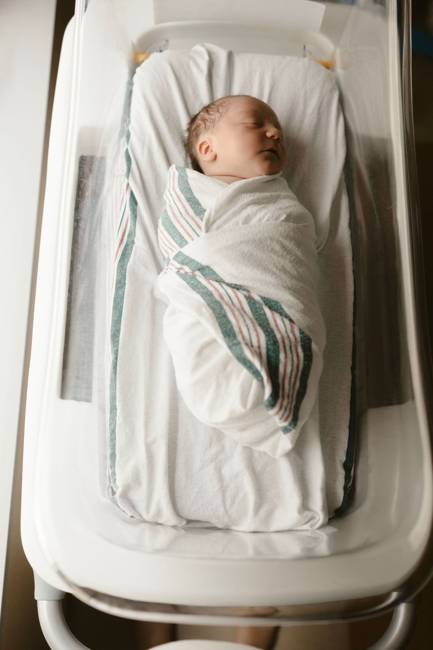 A sleeping infant wrapped in a white blanket with green stripes rests in a white bassinet viewed from different angles.