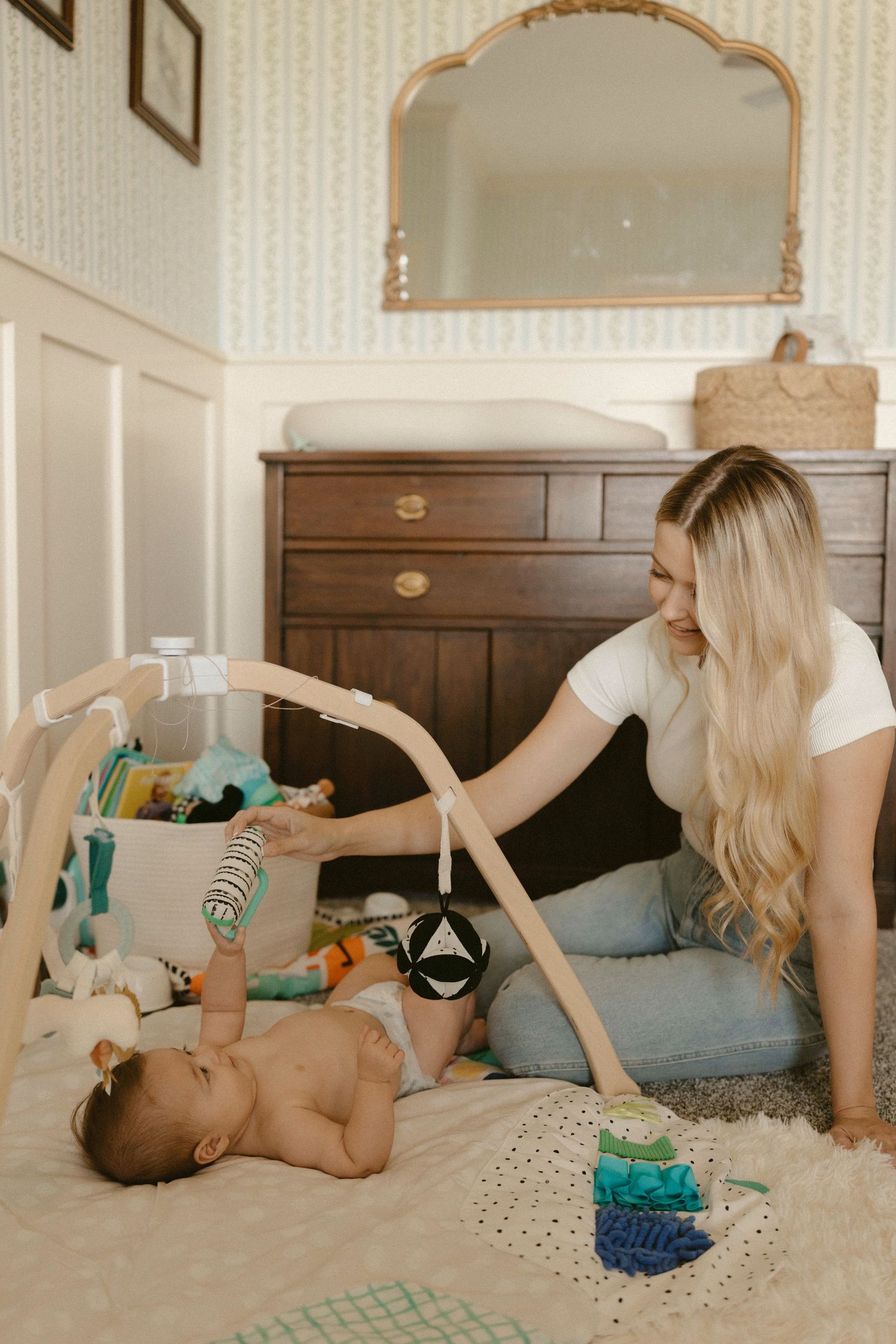 A baby lies on a plush carpet playing with a wooden activity gym while someone adjusts the hanging toys.