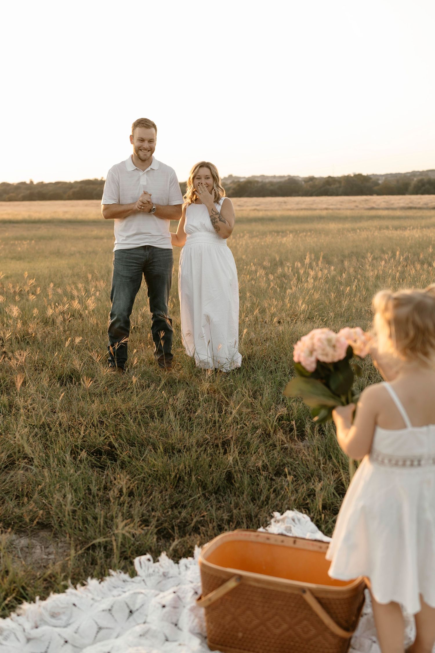 A couple in white outfits poses in a golden field at sunset while a child plays with a wagon nearby.