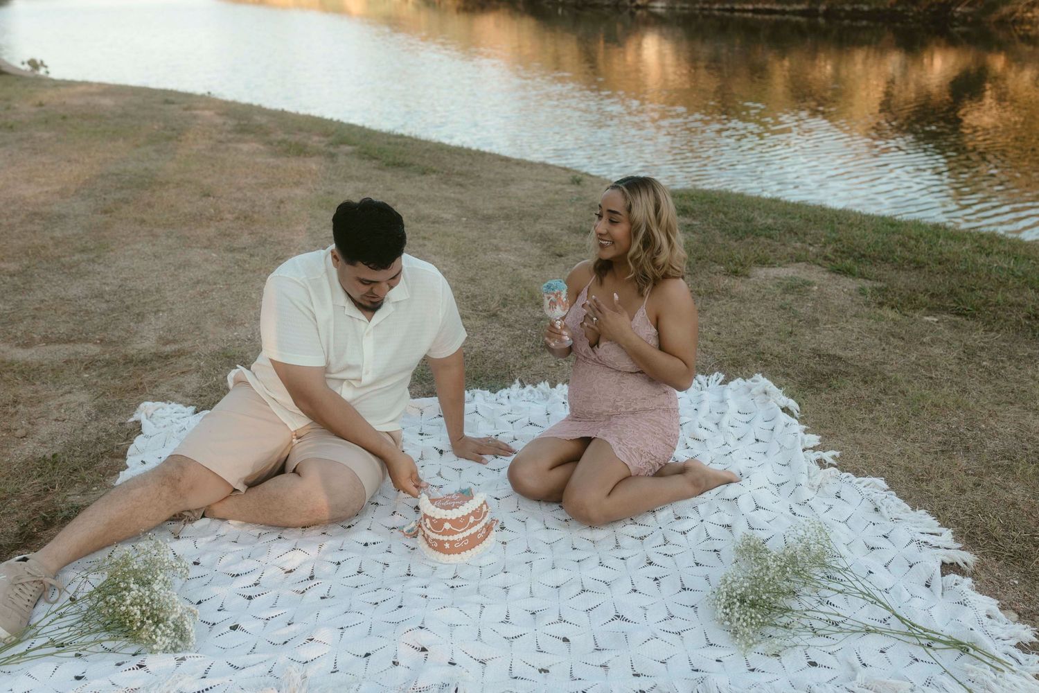 A couple sits on a blanket enjoying a picnic with wine glasses at sunset by a lake.