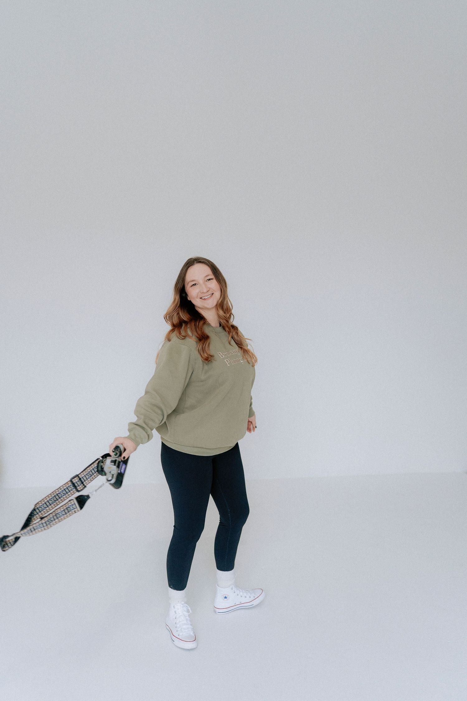 Person in casual attire wearing white sneakers and dark pants strikes playful poses against a light background with a camera in hand