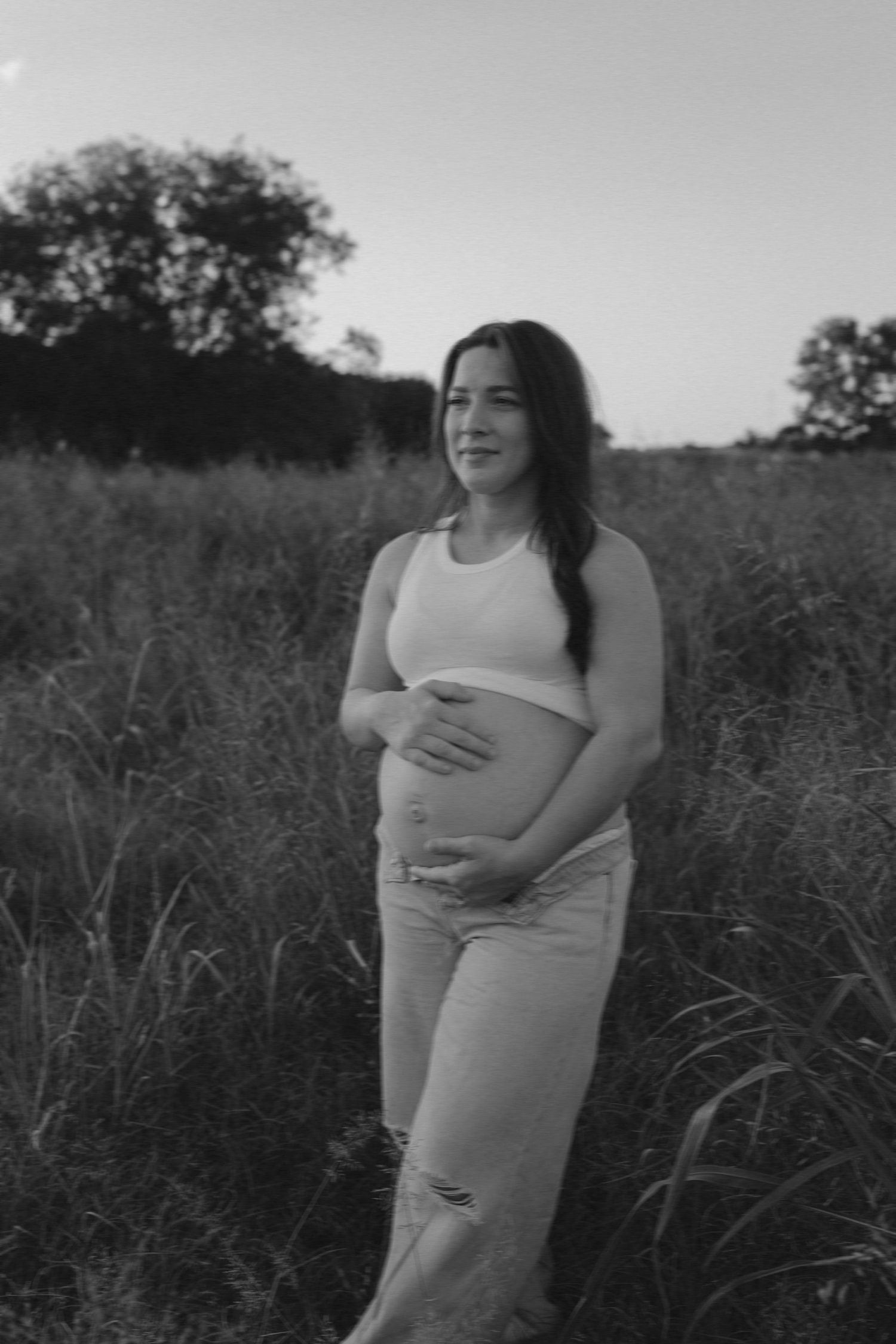 A series of black and white maternity photos show a pregnant woman in a white tank top embracing her belly in a field at dusk.