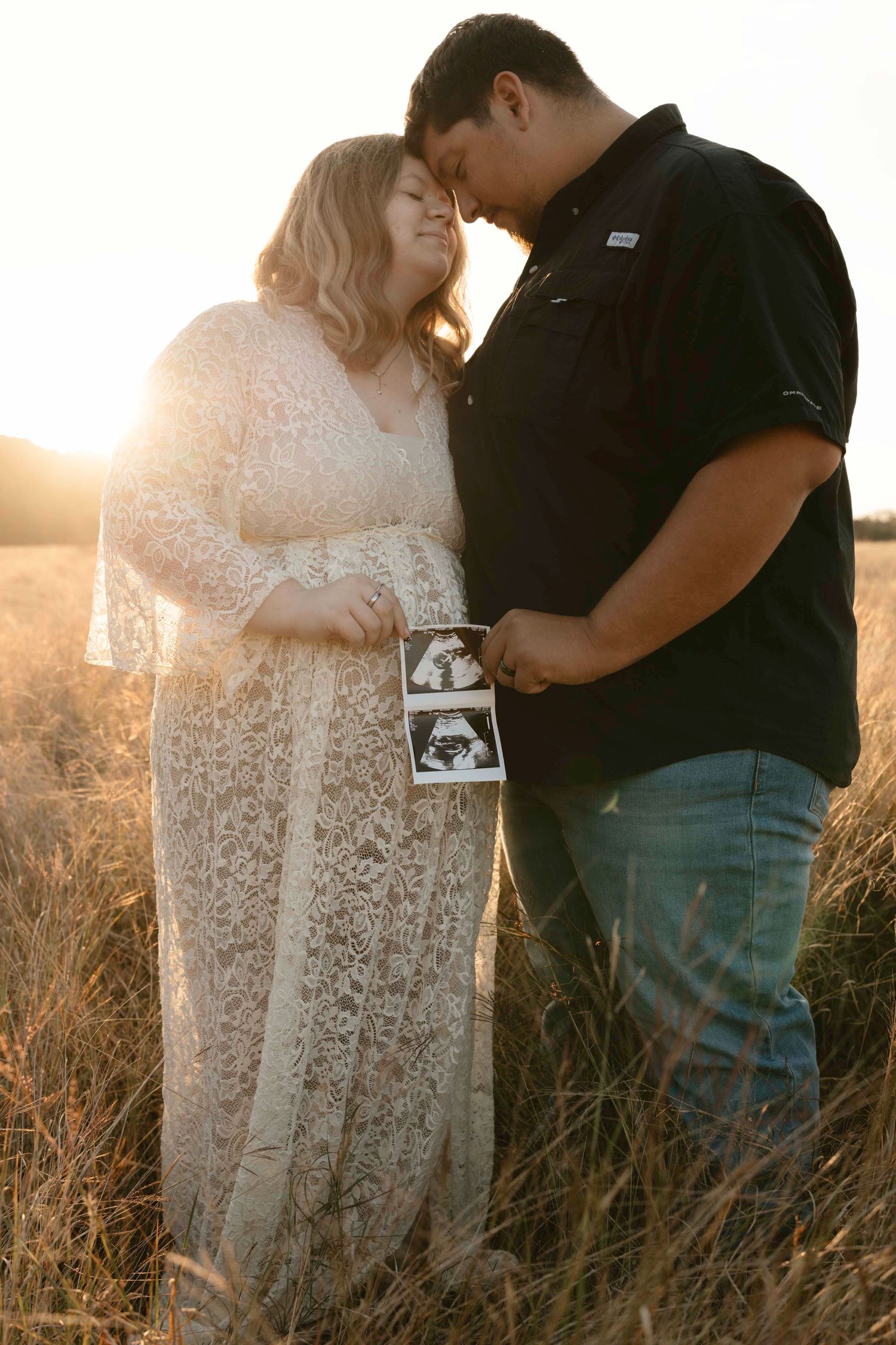 A couple embraces in a sunlit field of tall grass during golden hour, holding an ultrasound photo.