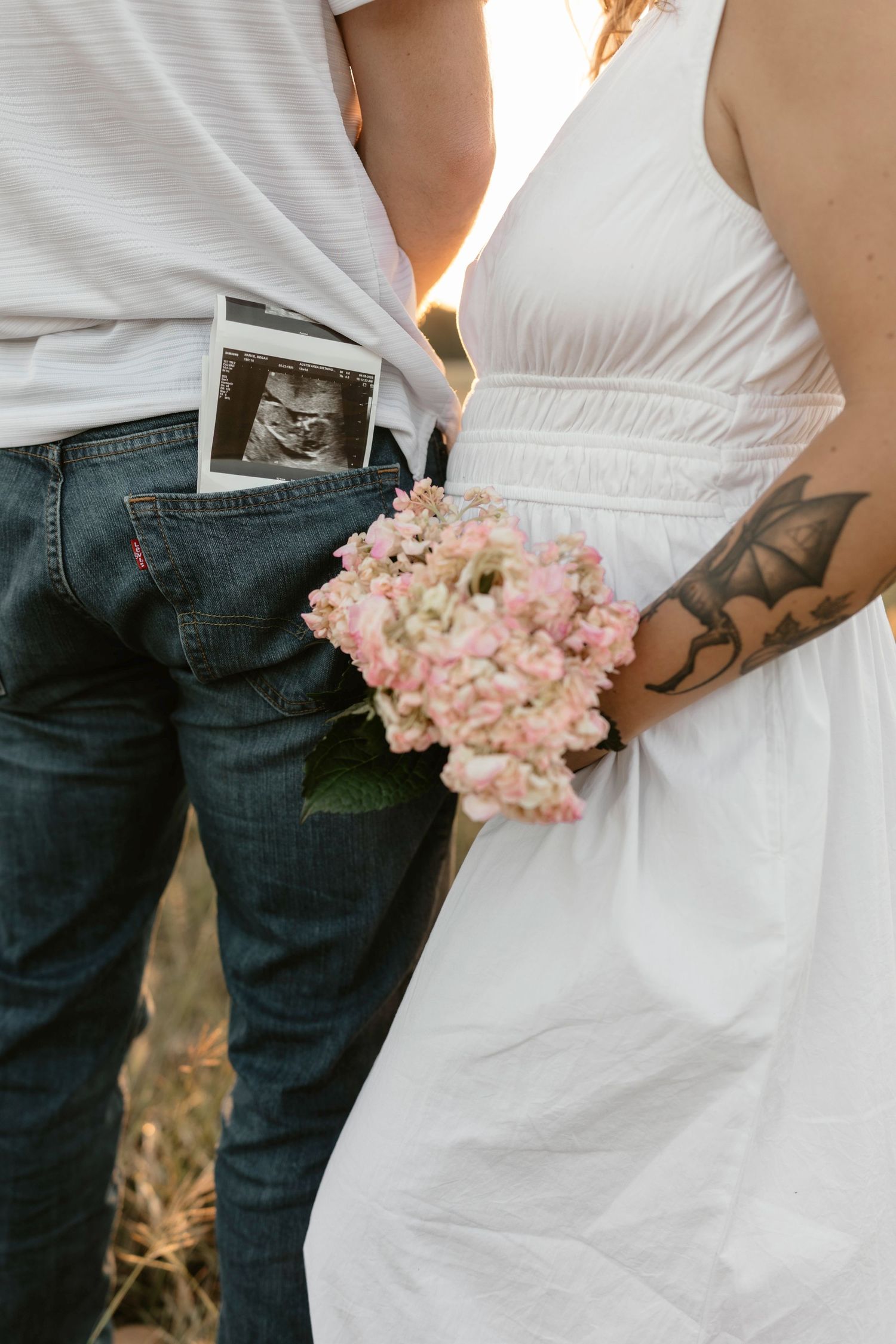 A couple in casual wedding attire, with denim jeans and white dress, holding pink hydrangea bouquet against sunset lighting.