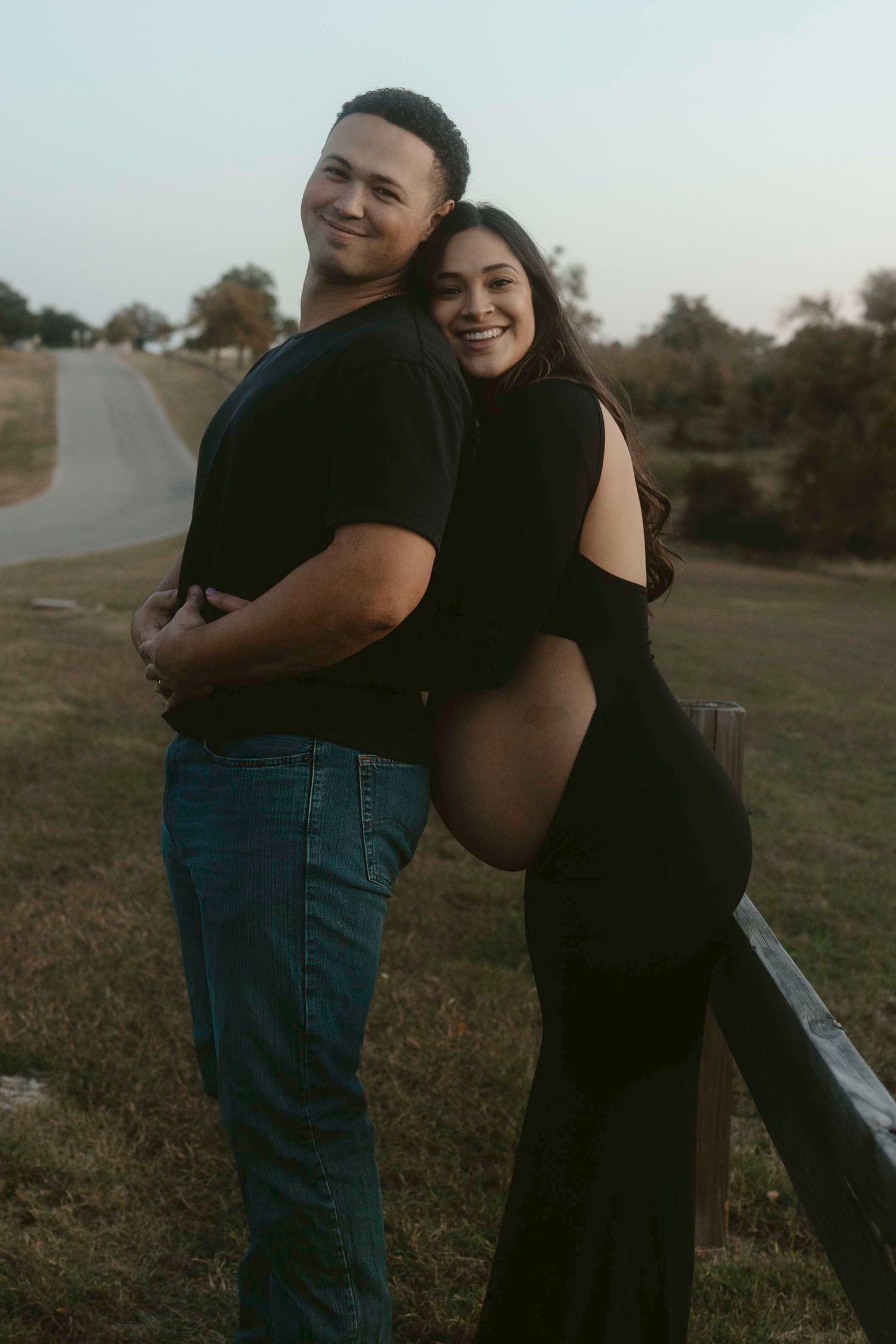 A couple embraces outdoors in a field during golden hour wearing black and denim clothing.