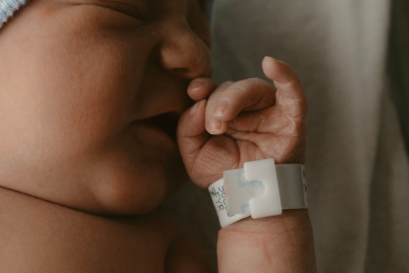 A newborn portrait of a tiny baby hand with a hospital ID bracelet resting near their face while sleeping.