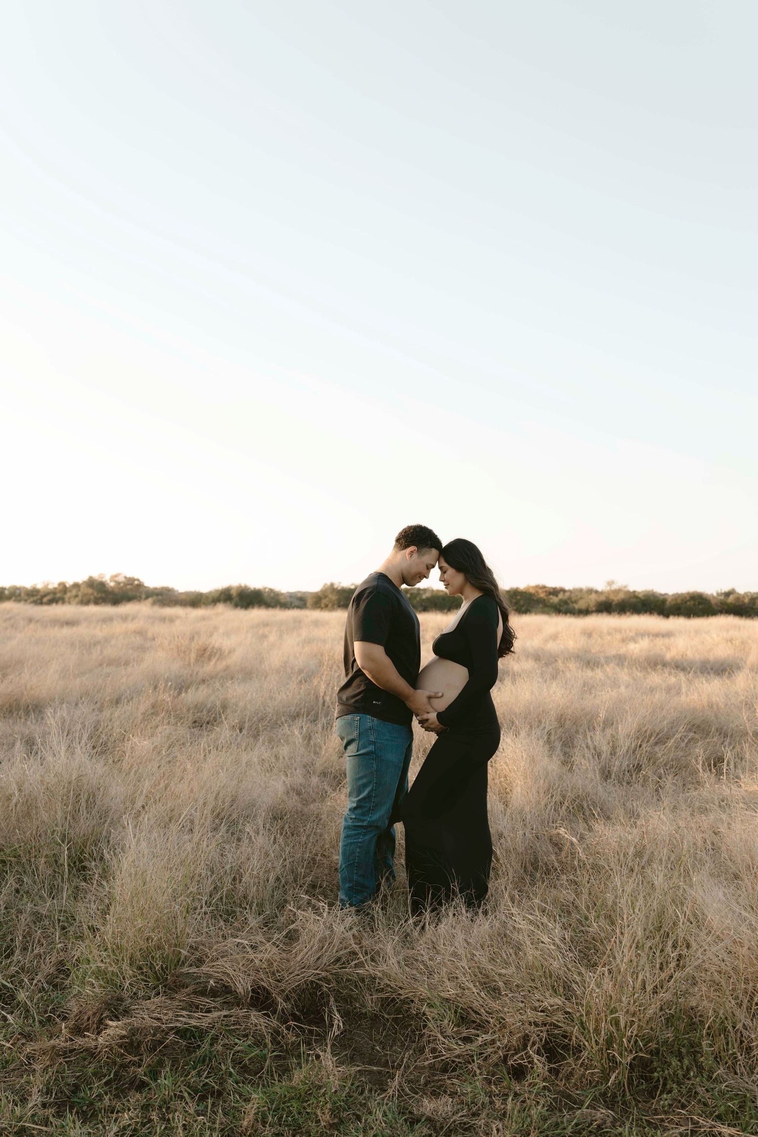 A maternity couple shares an intimate moment together while standing in a golden field at sunset.