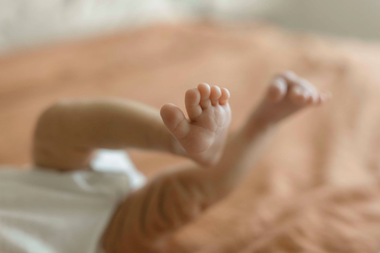 Tiny baby feet peek out from under soft beige bedding in a dreamy, blurred close-up photograph.