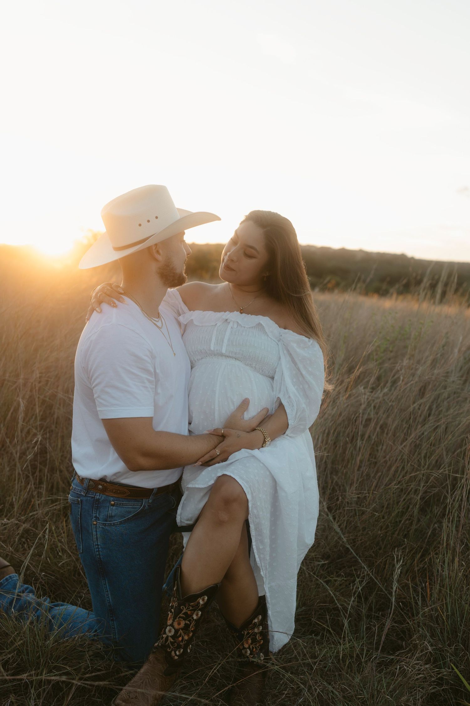A romantic maternity couple embraces in a golden wheat field at sunset wearing white and denim.