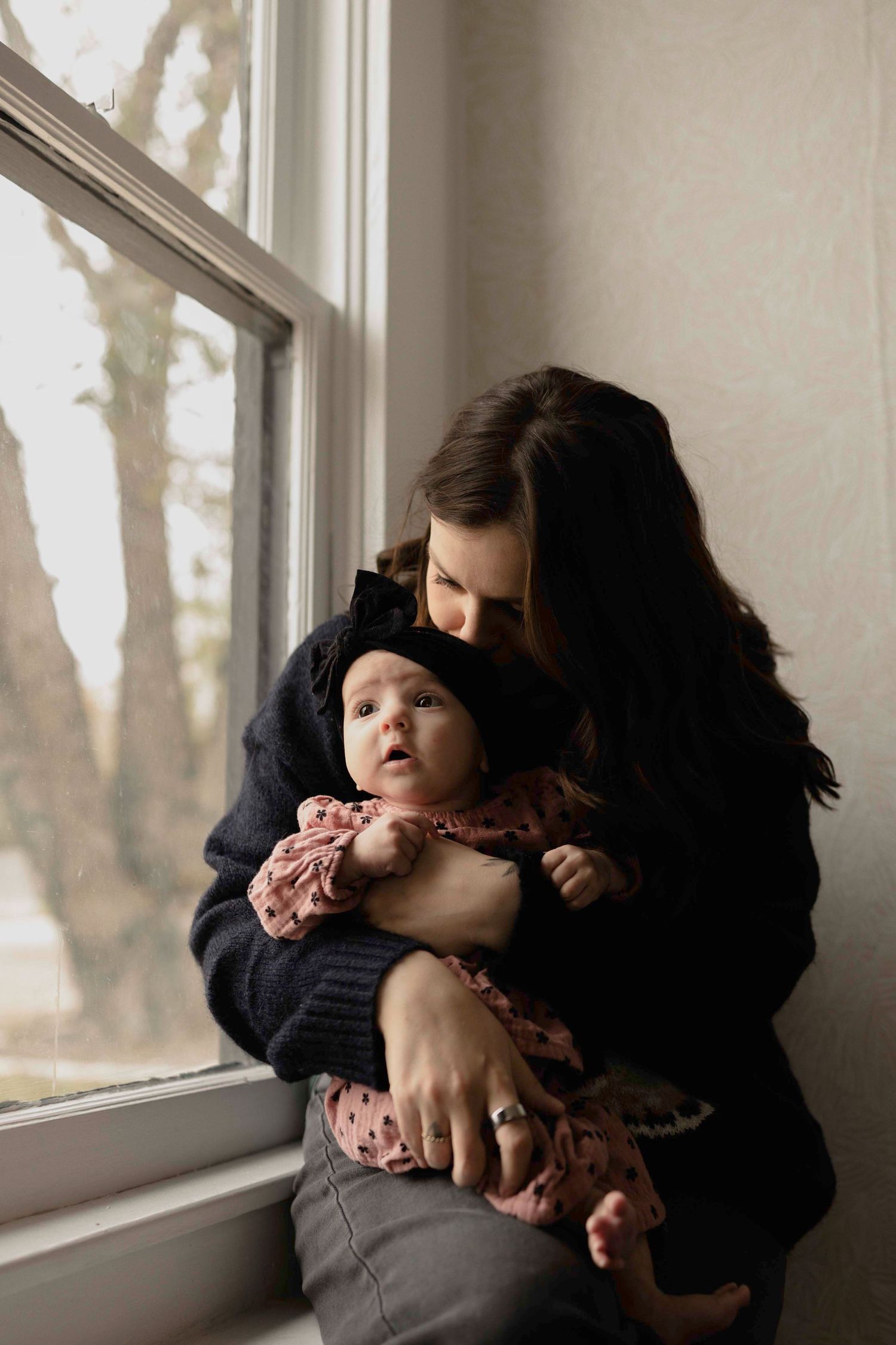 A tender moment by a window as a parent in black sweater embraces their small baby wearing pink and navy clothes.