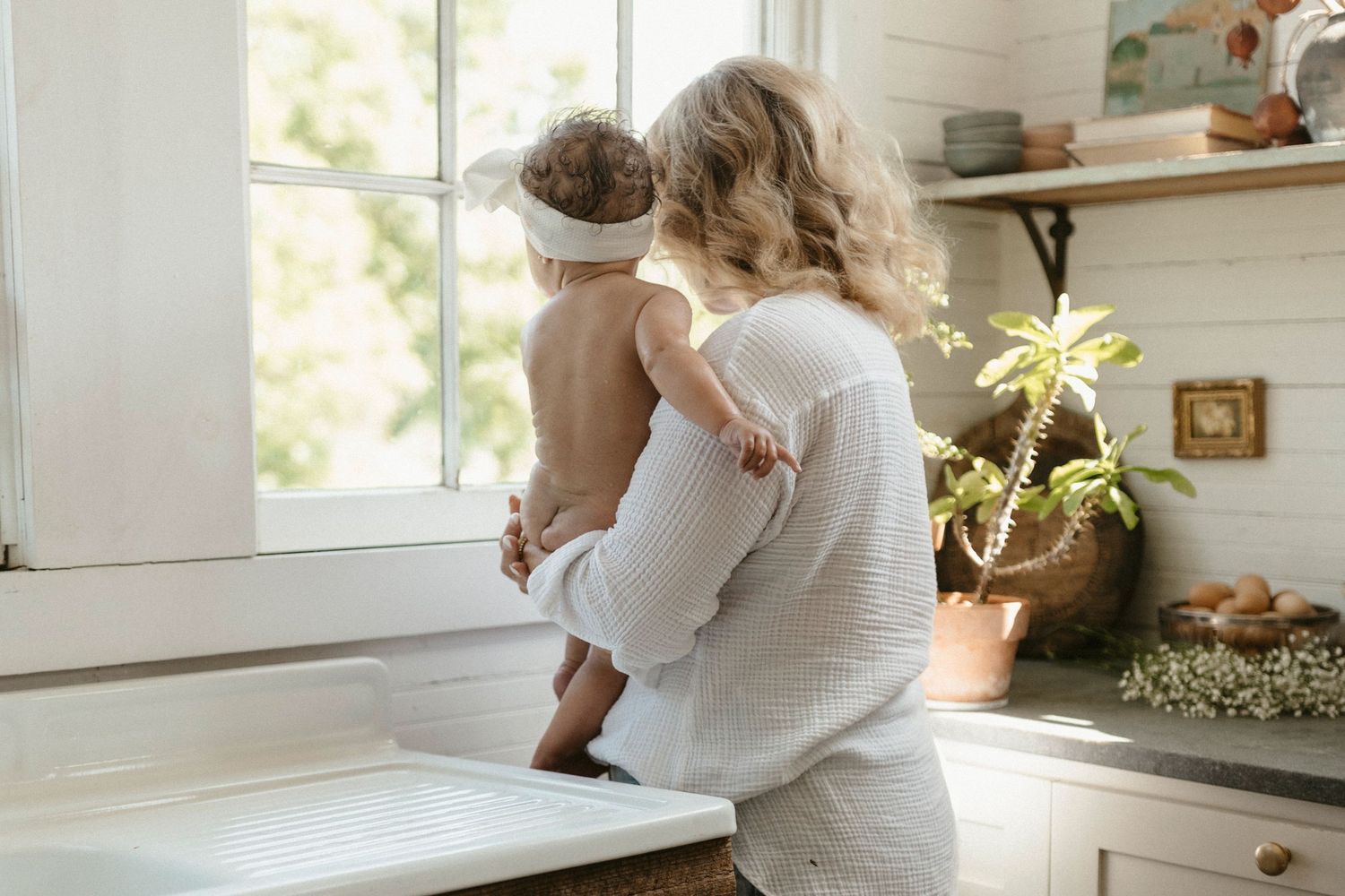 A motherhood portrait in a flowing white dress, while she holds her child while preparing food in a bright, airy kitchen.