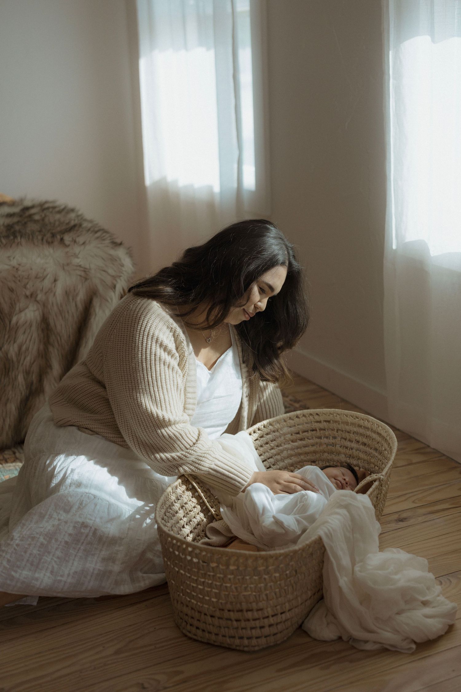 Minimalist newborn photo of mother in white dress and cream cardigan sitting beside woven bassinet with newborn.