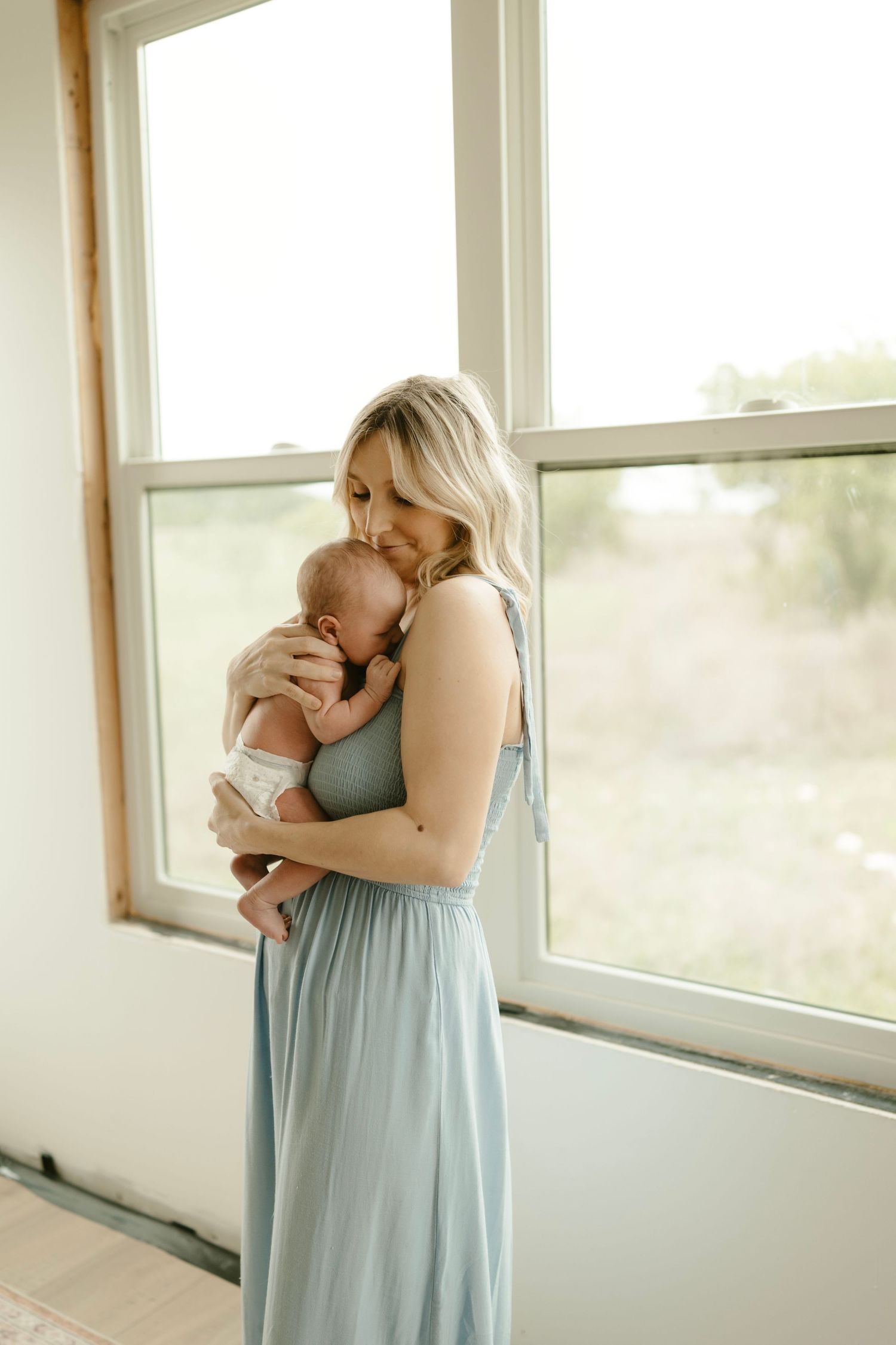 Newborn photo session with mother in blue dress holding baby near sunlit window.