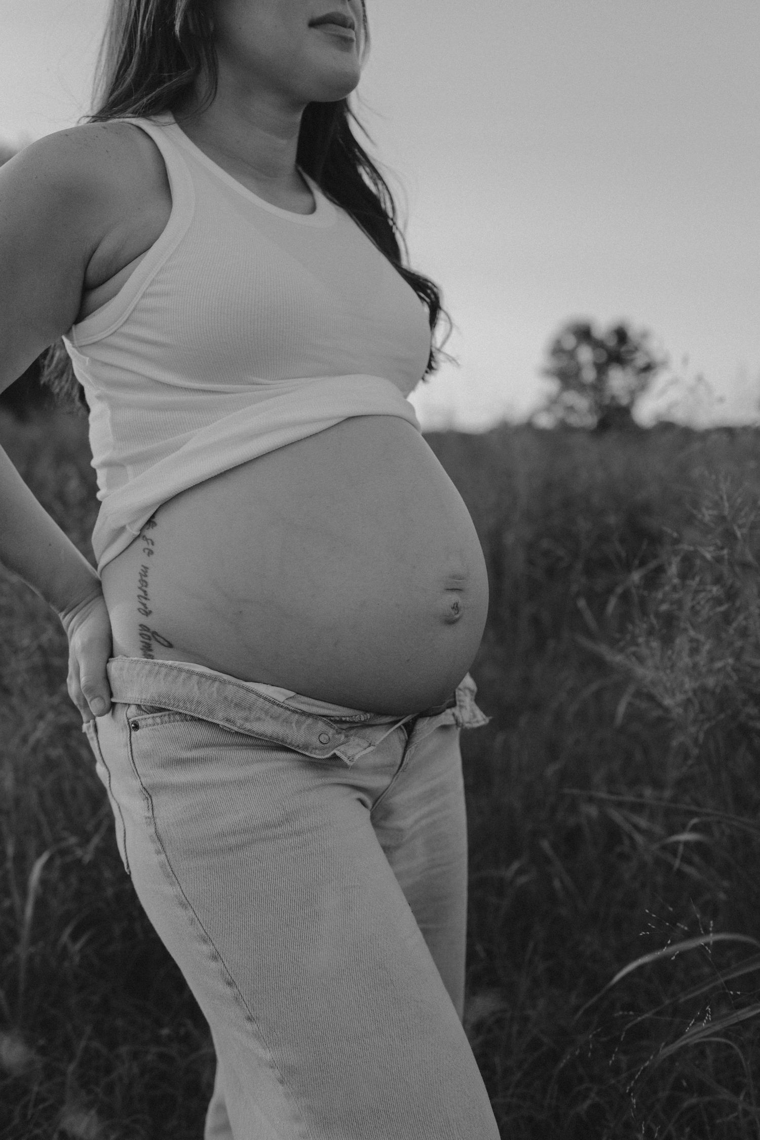 Black and white artistic maternity photo showing a pregnant belly in a natural outdoor setting with tall grass.
