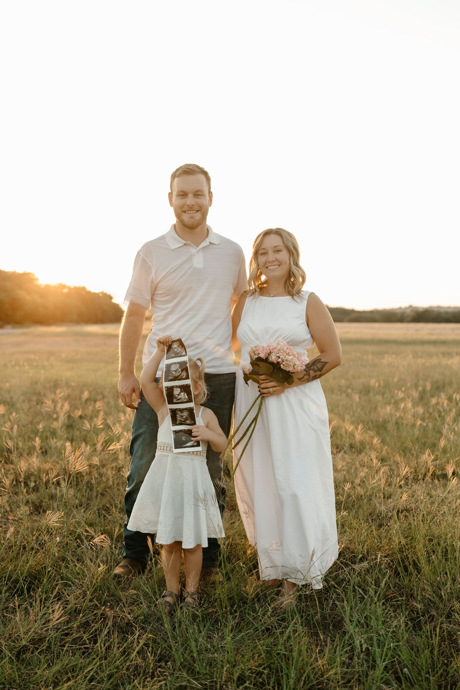 A couple poses in a golden field at sunset while holding ultrasound photos and a bouquet of flowers.