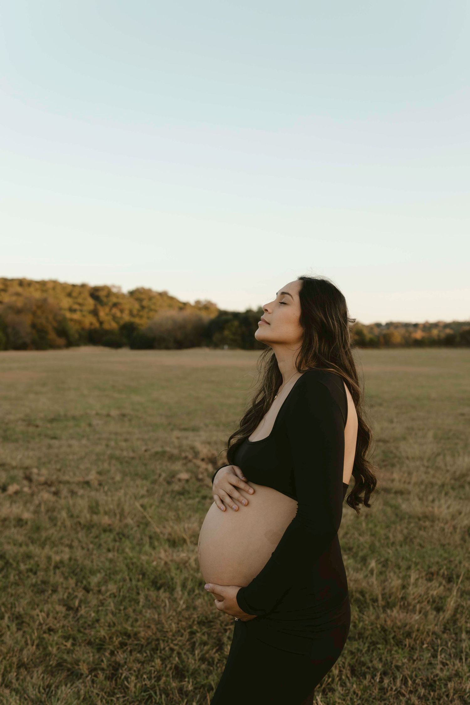 Expectant mother in black dress cradles her baby bump while standing in a golden field at sunset.