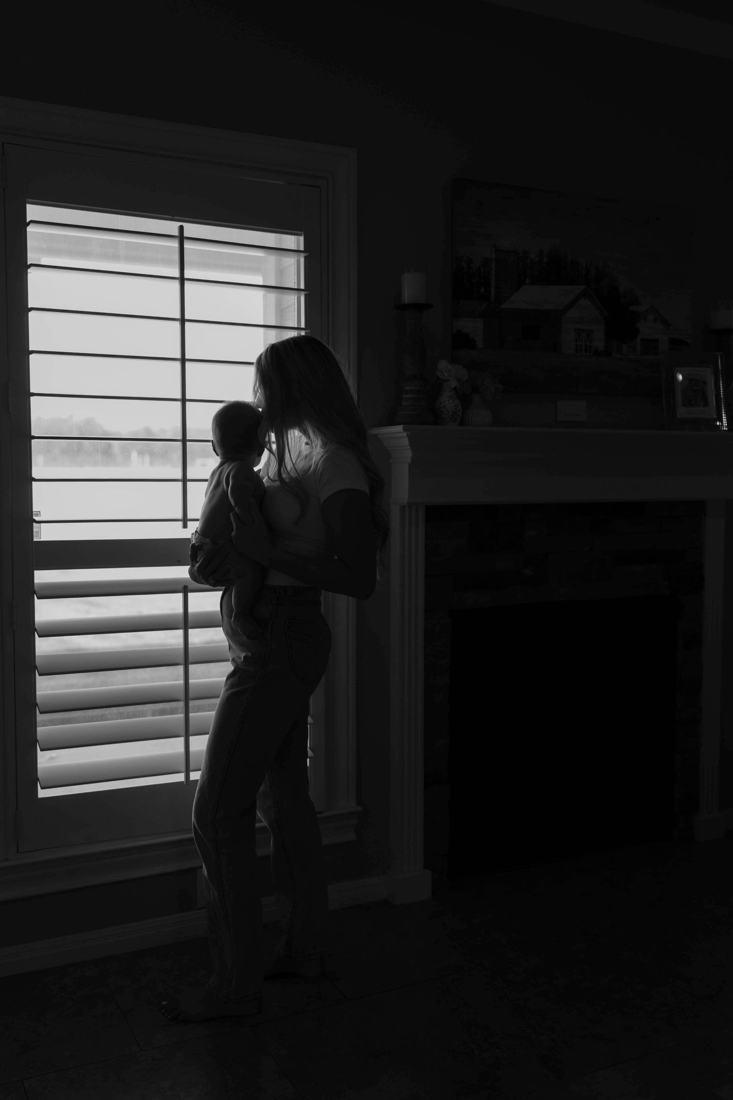 Motherhood photography of a mom holding her baby while looking out a window inside their home.