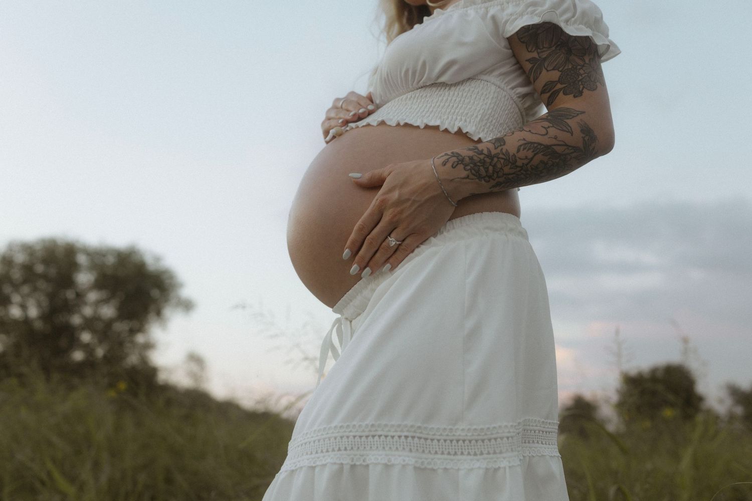 A pregnant woman in a white dress standing in a natural outdoor setting at sunset.