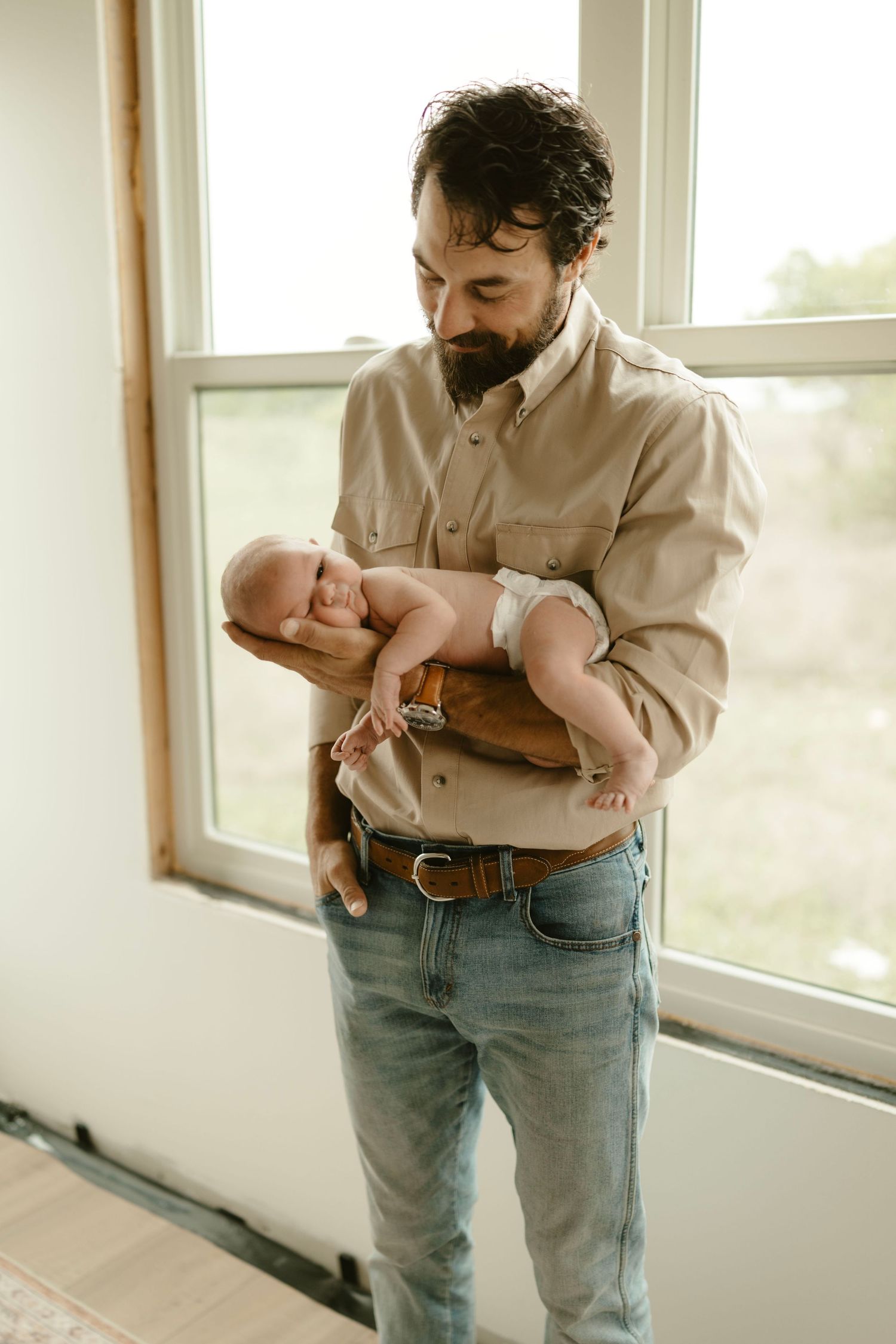 Fun newborn photo of dad in denim holding his infant by bright window.
