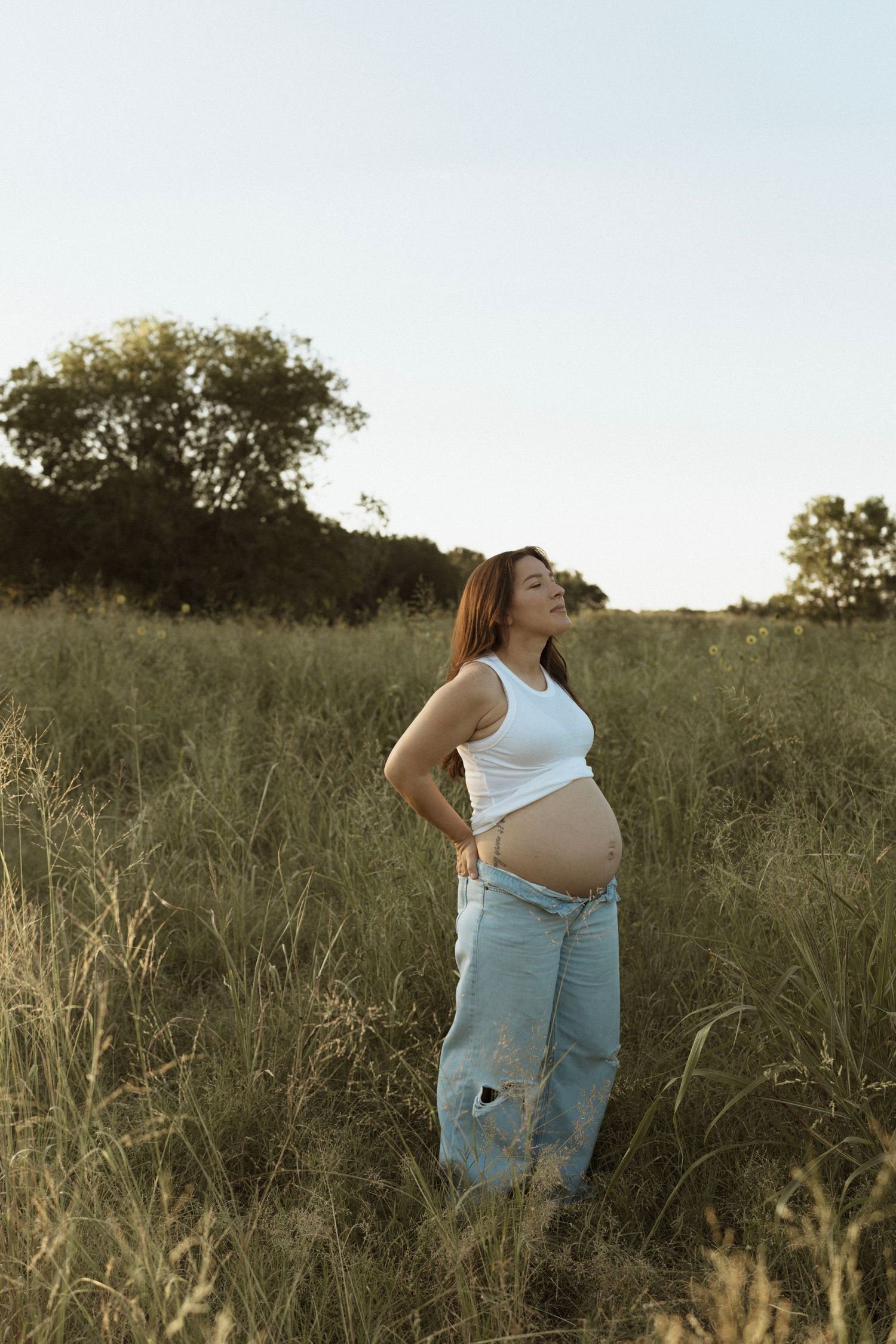 A pregnant person in denim overalls stands in a sunlit meadow at golden hour among tall grass and wildflowers.