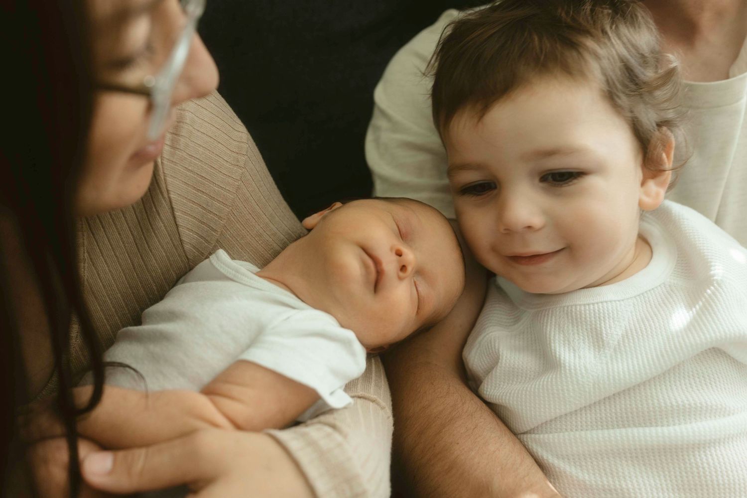Two children in white sweaters share a tender moment while resting together with an adult nearby.
