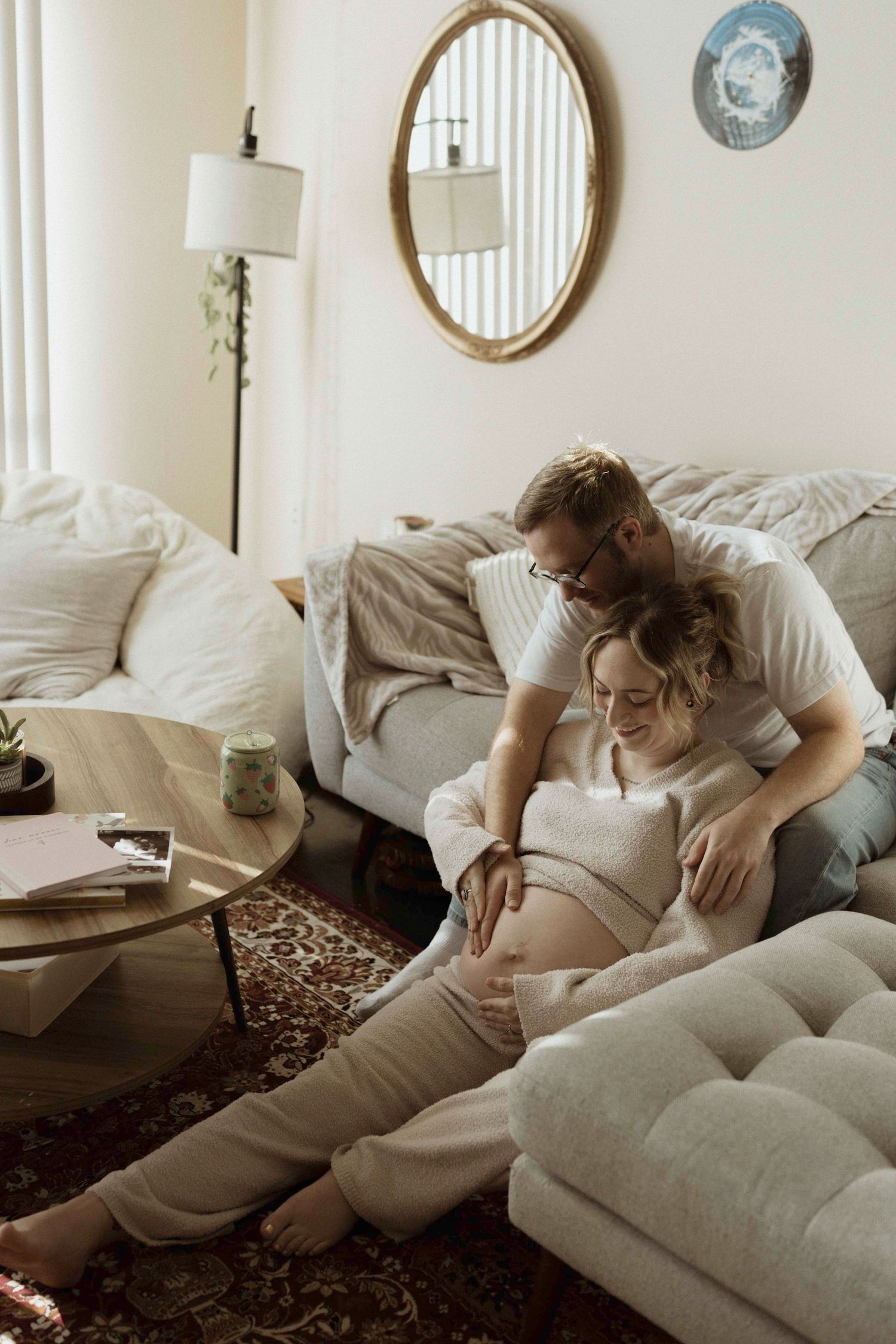 A couple lounges intimately together on a white couch in a sunlit room with circular mirrors on the wall, looking down at a pregnant belly.