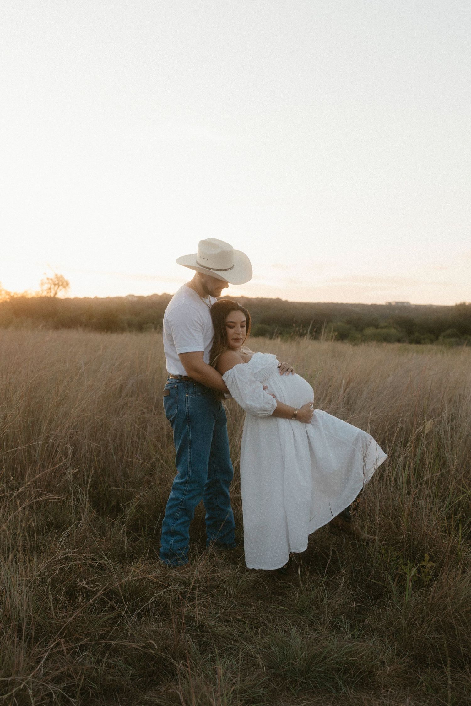 A couple in western attire embrace in a golden field at sunset, with her wearing a flowing white dress, holding a baby bump.
