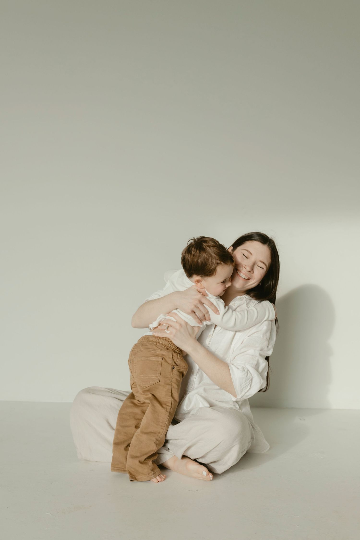 Minimalist motherhood photography showing a mom holding and cuddling her son in a white studio setting.