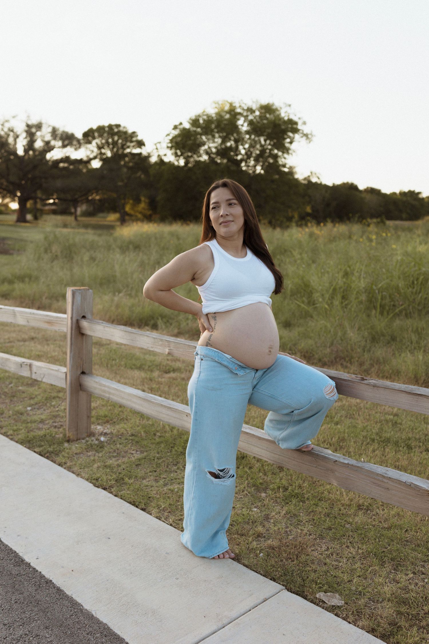 A pregnant mom in light blue pants strikes various poses along a concrete path near a wooden fence.