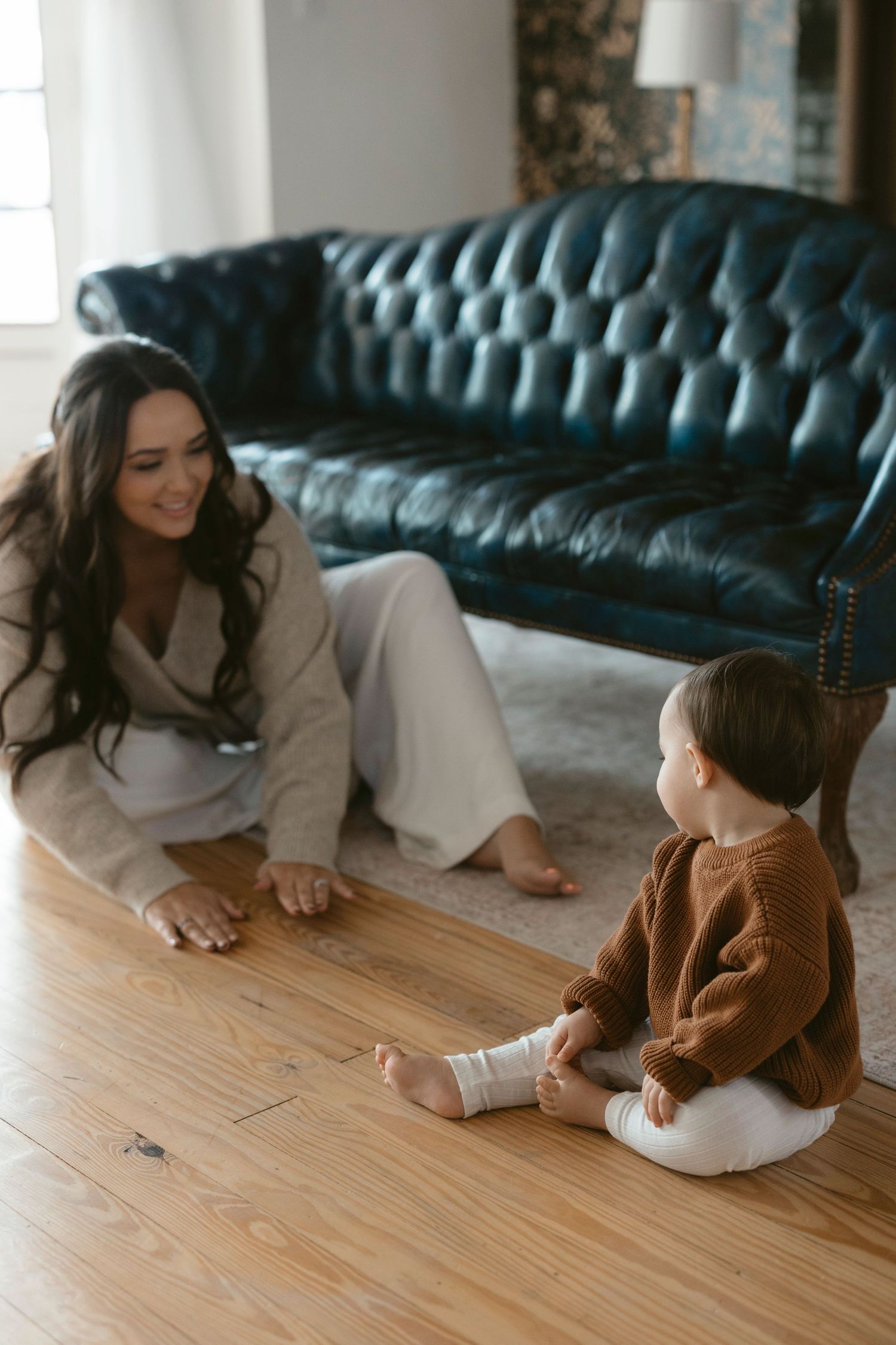 Motherhood photo session in Austin, Texas showing parent and child sharing playful moment on wooden floor near tufted leather couch.