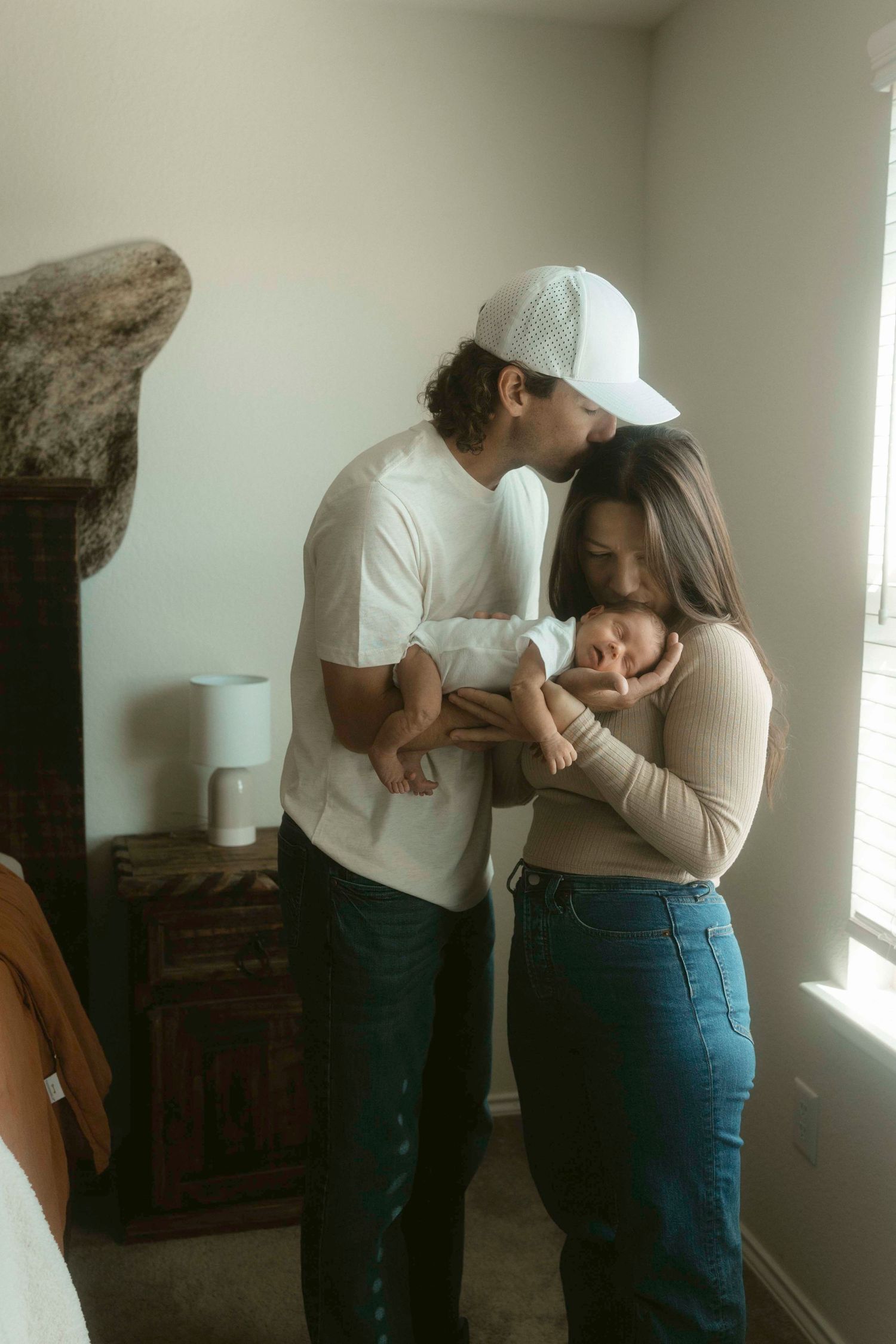 A couple embraces in a softly lit room against a white wall with minimalist decor, holding a newborn baby.
