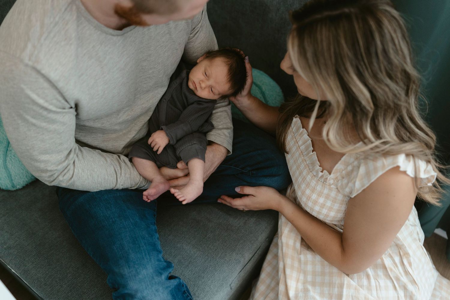 A couple sits close together on a couch sharing an intimate moment holding a newborn baby.