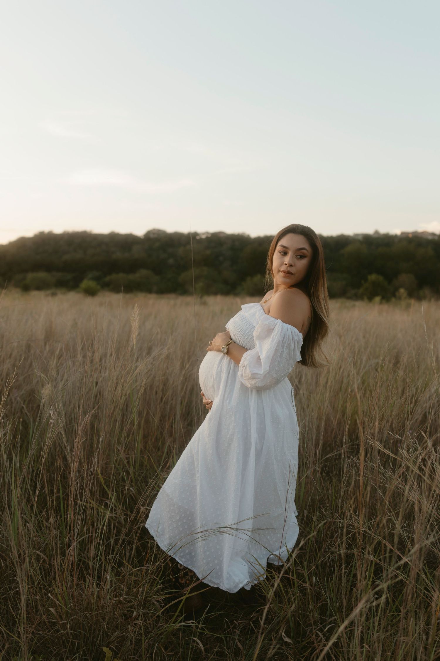 A maternity portrait a flowing white dress poses gracefully in a golden field at sunset, creating an ethereal scene.