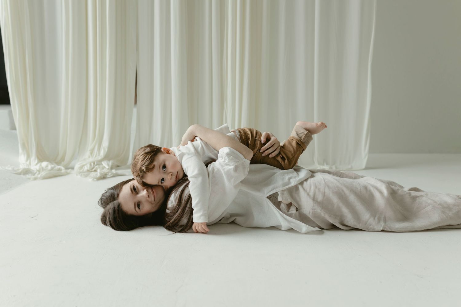 Two people in beige loungewear lie on white floor near curtains in soft natural light.