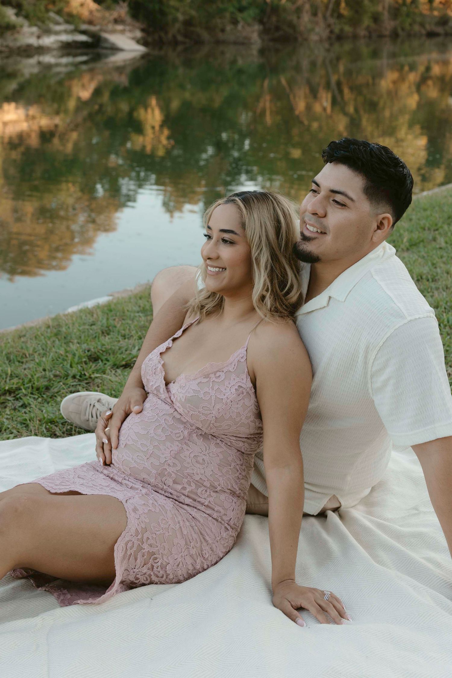 A couple in pink and white outfits relaxes by a scenic pond during a romantic photoshoot session.