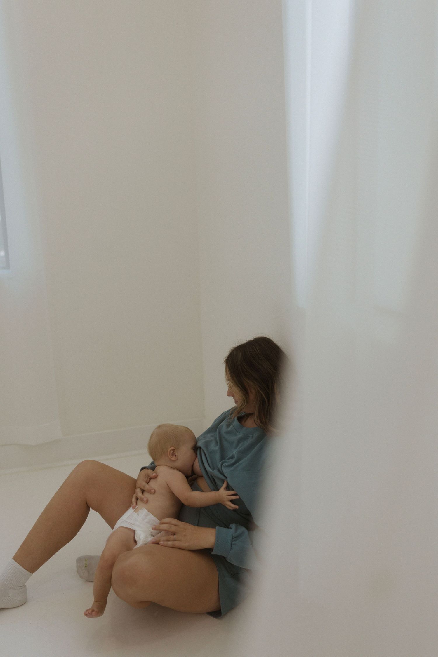A mom sits against a white wall in casual clothing while breastfeeding a baby wearing a diaper.