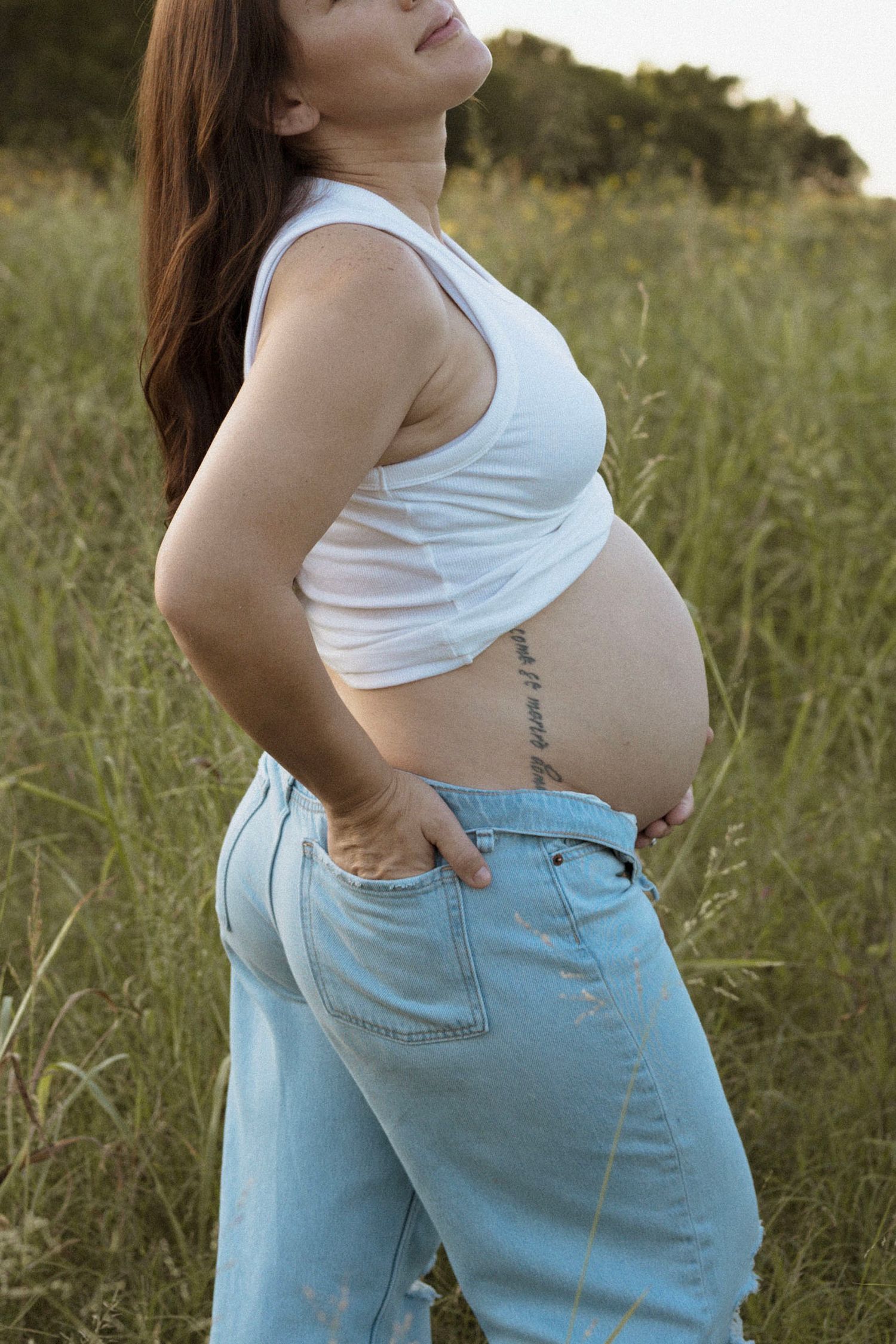 A pregnant mom wearing a white tank top and light blue jeans stands in a grassy outdoor field during golden hour.