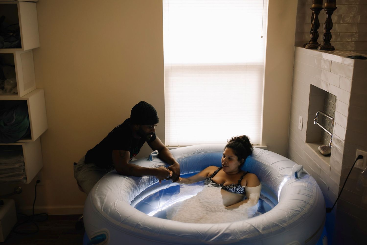 Two people relax in a home birth pool filled with water during labor preparation in a dimly lit room.