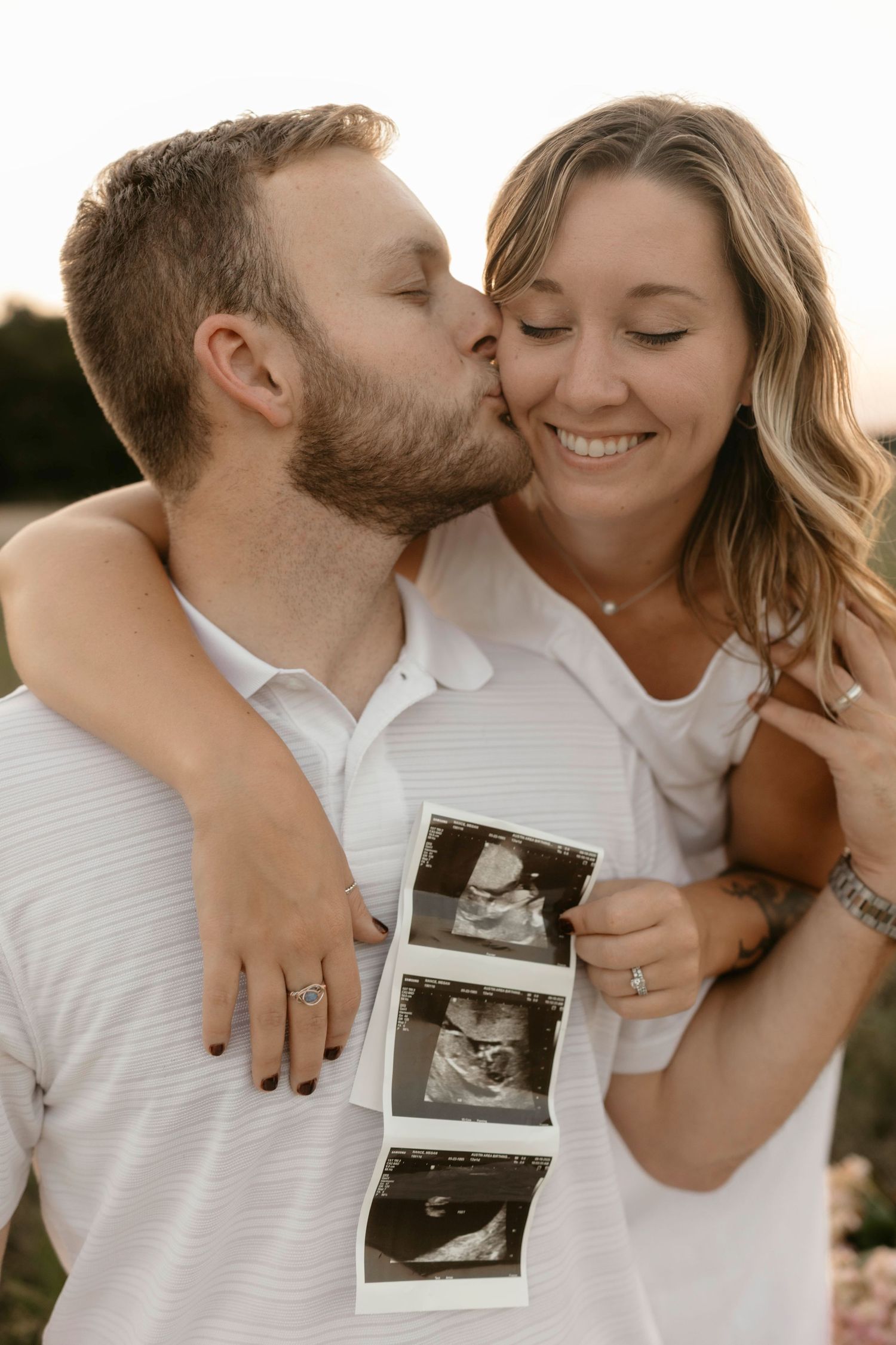 A romantic couple shares an intimate moment while holding ultrasound photos in warm evening light.