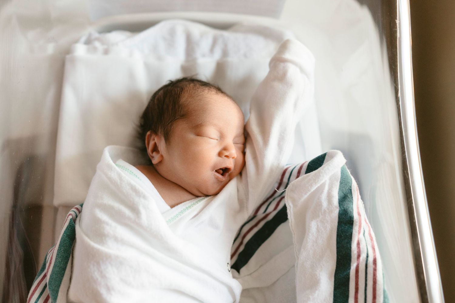 A sleeping newborn wrapped in white swaddle blanket with teal stripes rests peacefully near a window.