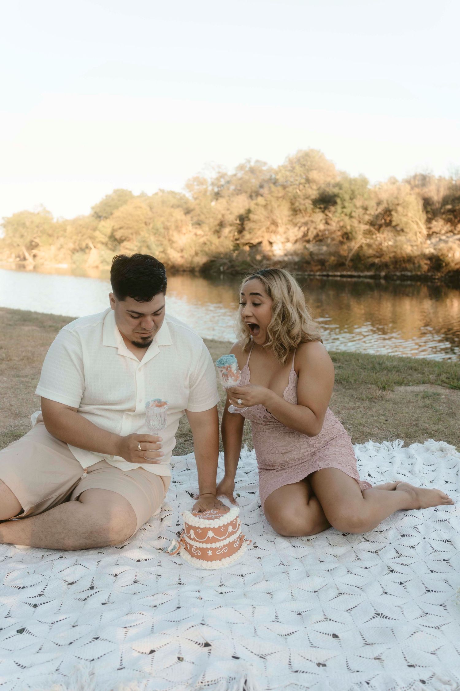 A couple sits on a white blanket by a lake enjoying a romantic picnic with cake and drinks at sunset.