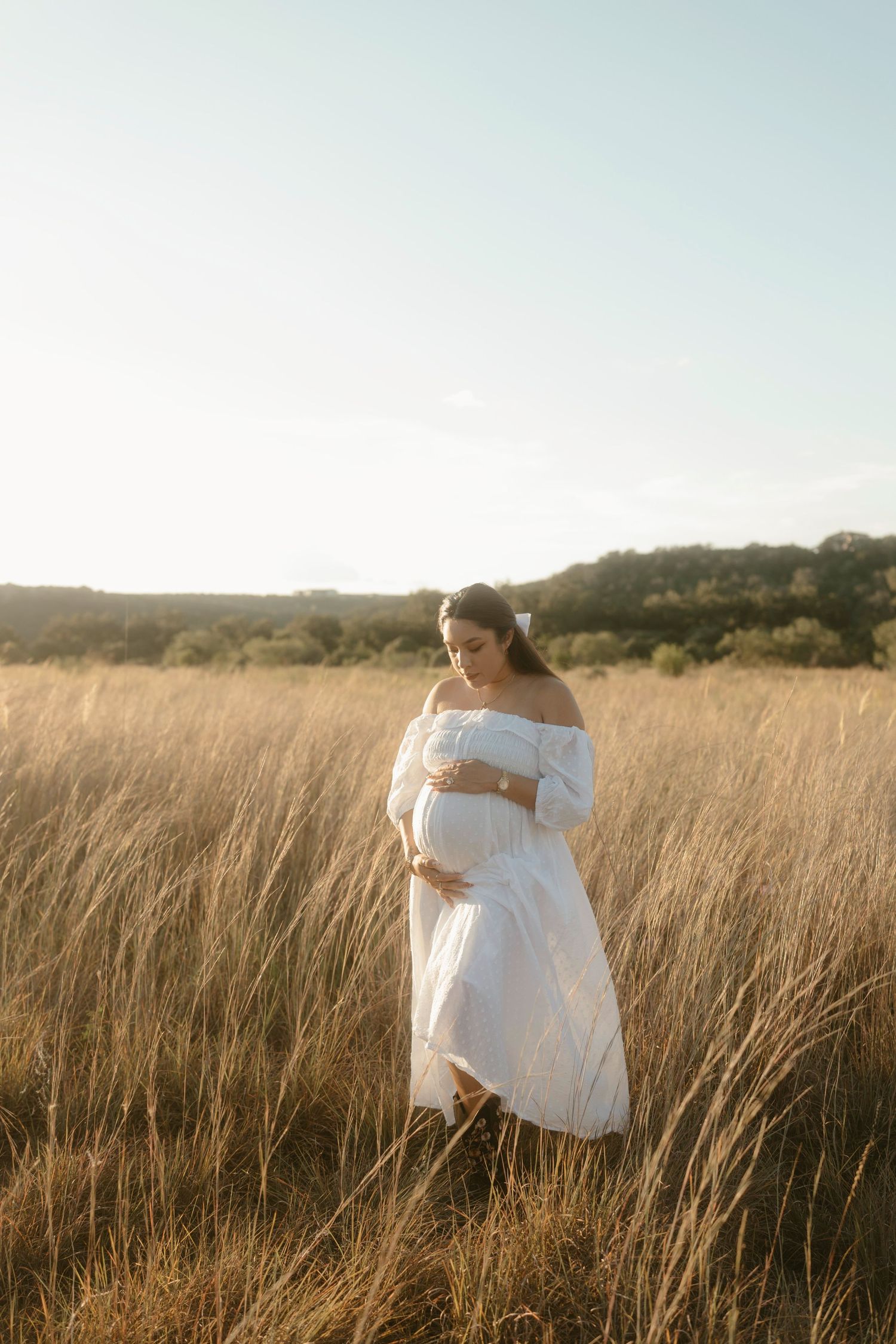 An expectant mother in a flowing white dress stands peacefully among tall grass at golden hour.