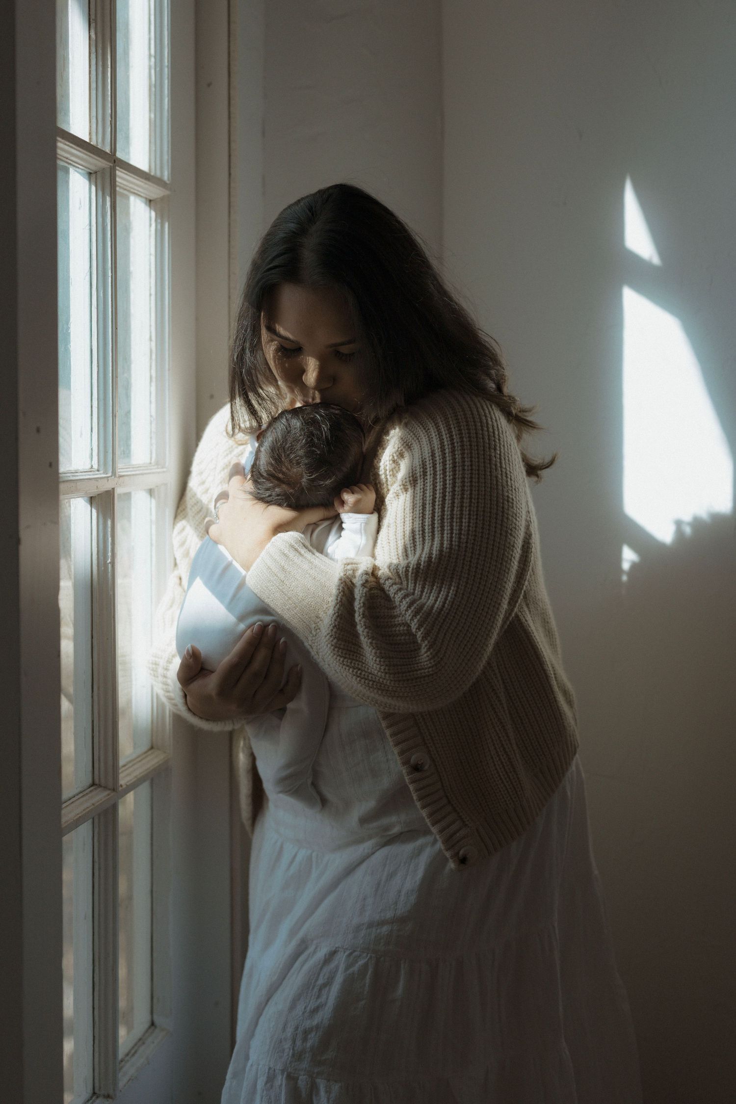 Moody window portraits showing a person in a white dress and sweater looking kissing her newborn baby