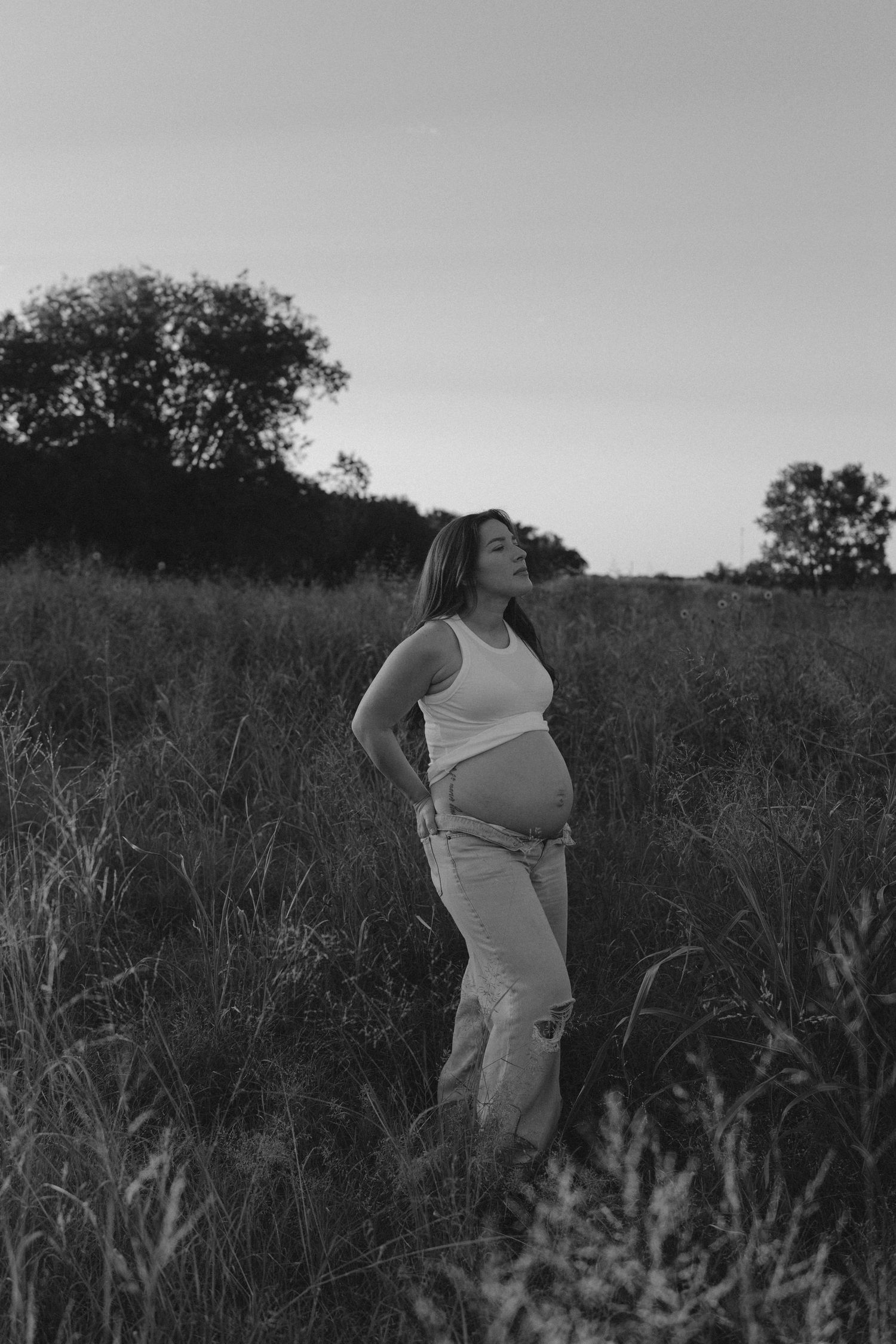 A black and white portrait of a pregnant person standing in a field of tall grass at sunset.
